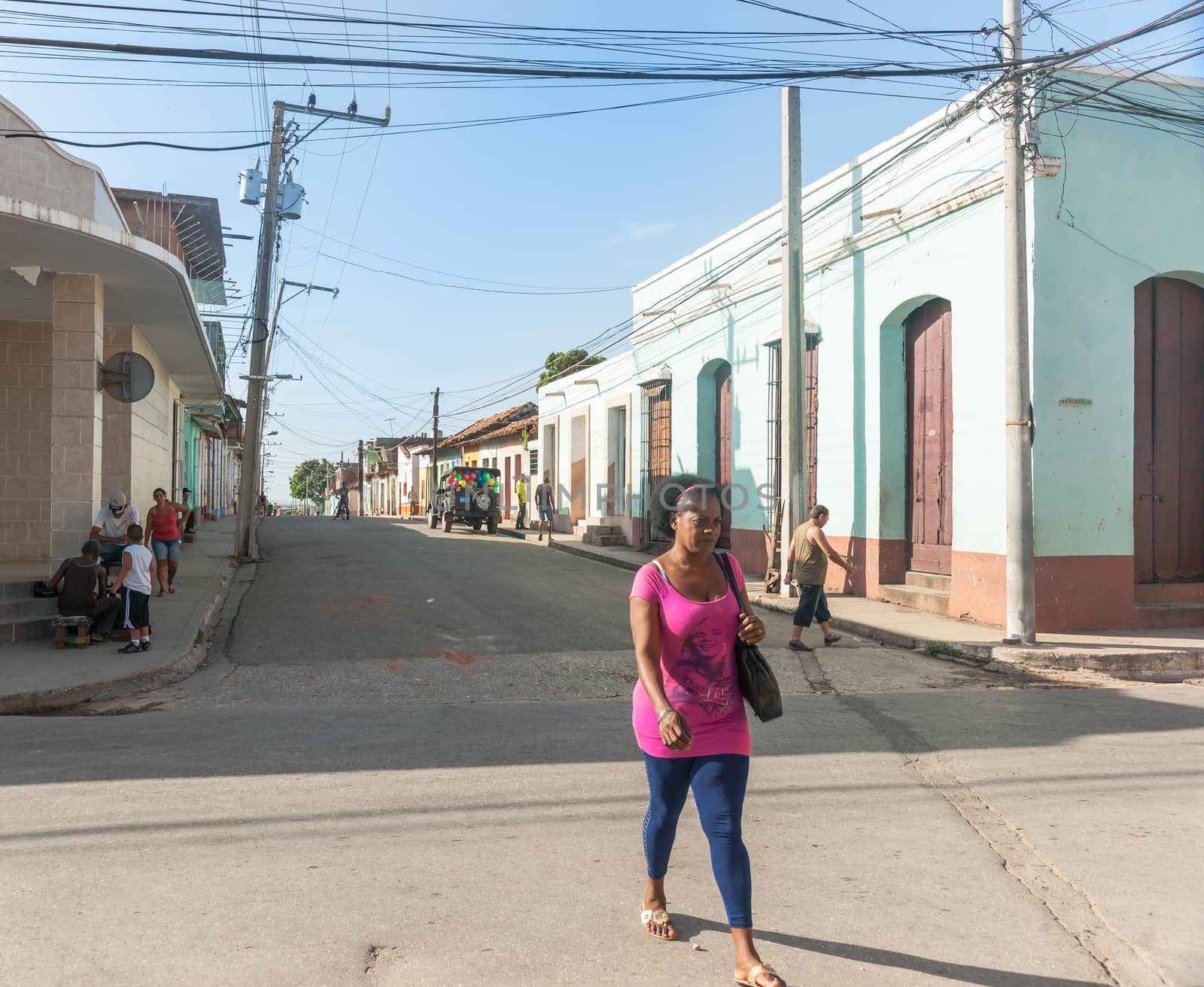 Trinidad, Cuba - July 2, 2012: Woman in pink top stands out as she walks along street lined by colonial style in Cuban town where others are sitting on corner