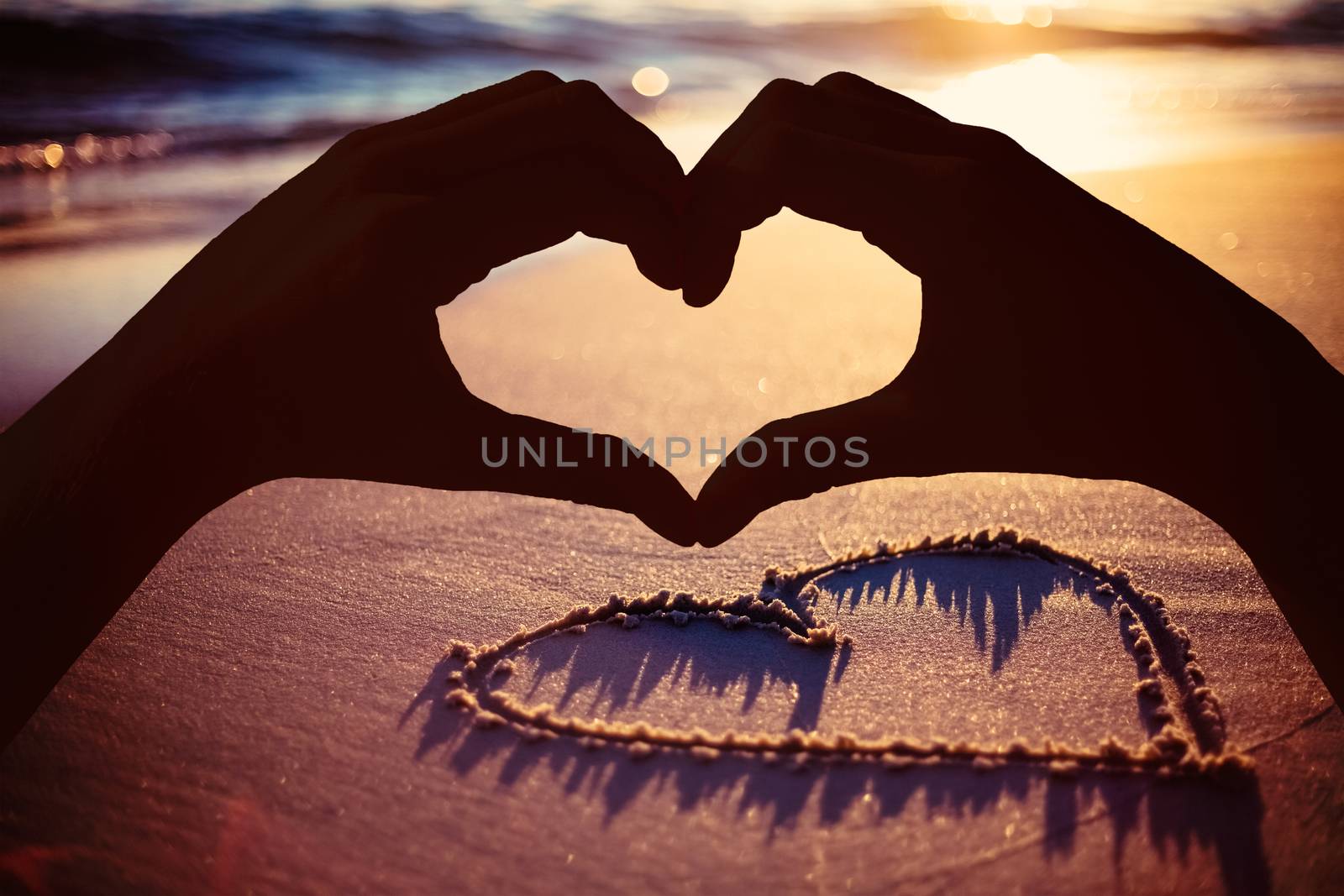 Hands making heart shape on the beach against one heart drawn in the sand 