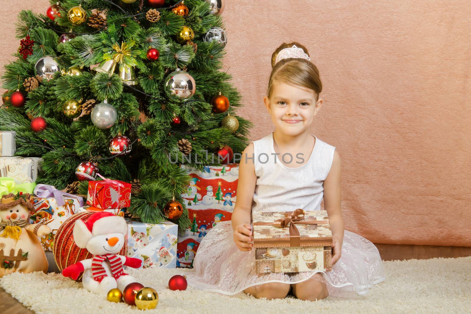 Five-year girl in a New Year's dress sitting on a mat at the Christmas tree