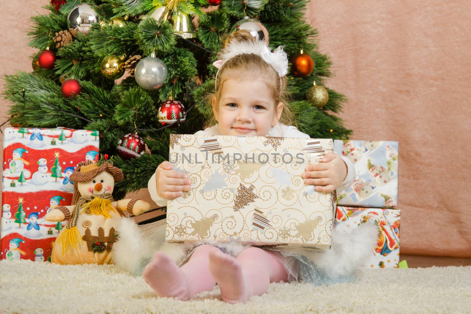 The three-year girl in a New Year's dress sitting on a mat at the Christmas tree