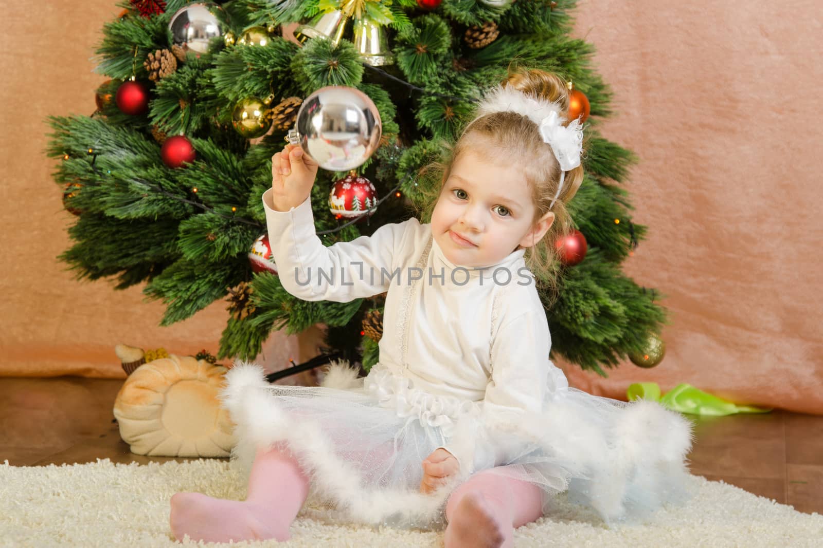 The three-year girl in a New Year's dress sitting on a mat at the Christmas tree