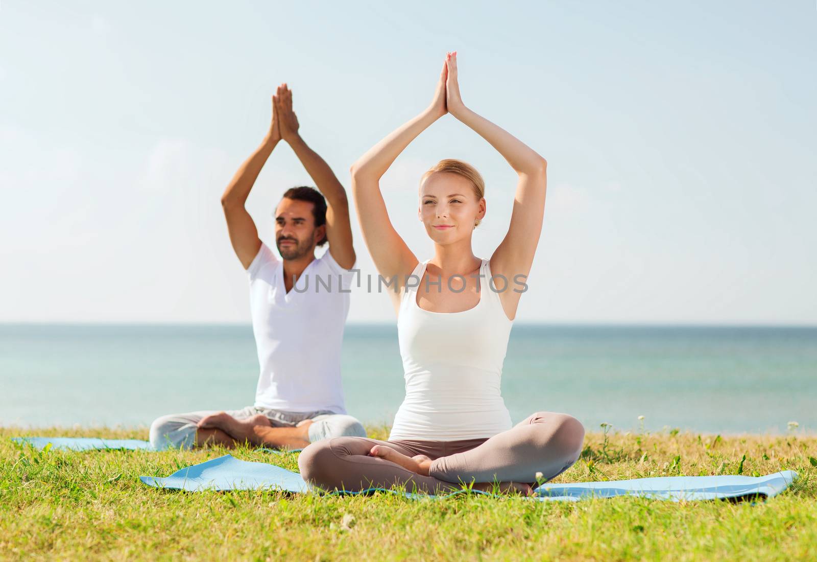 fitness, sport, friendship and lifestyle concept - smiling couple making yoga exercises sitting on mats outdoors