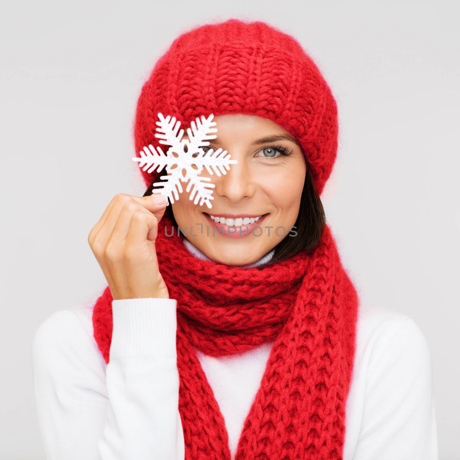 happiness, winter holidays, christmas and people concept - smiling young woman in red hat, scarf and mittens covering one eye with snowflake decoration over gray background