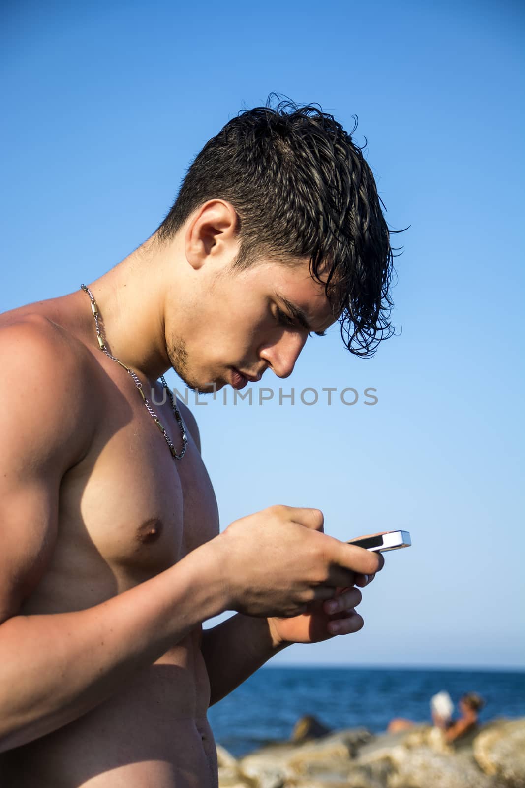 Shirtless Young Handsome Man Busy with his Mobile Phone While Standing at the Beach Boulders next to Sea or Ocean