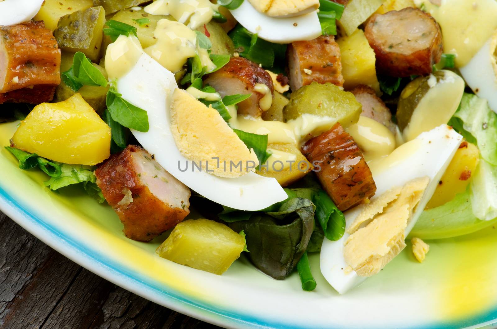 Homemade Delicious Potato and Smoked Sausage Salad with Gherkins, Lettuce, Boiled Eggs and Mustard Sauce on Yellow and Green Plate closeup