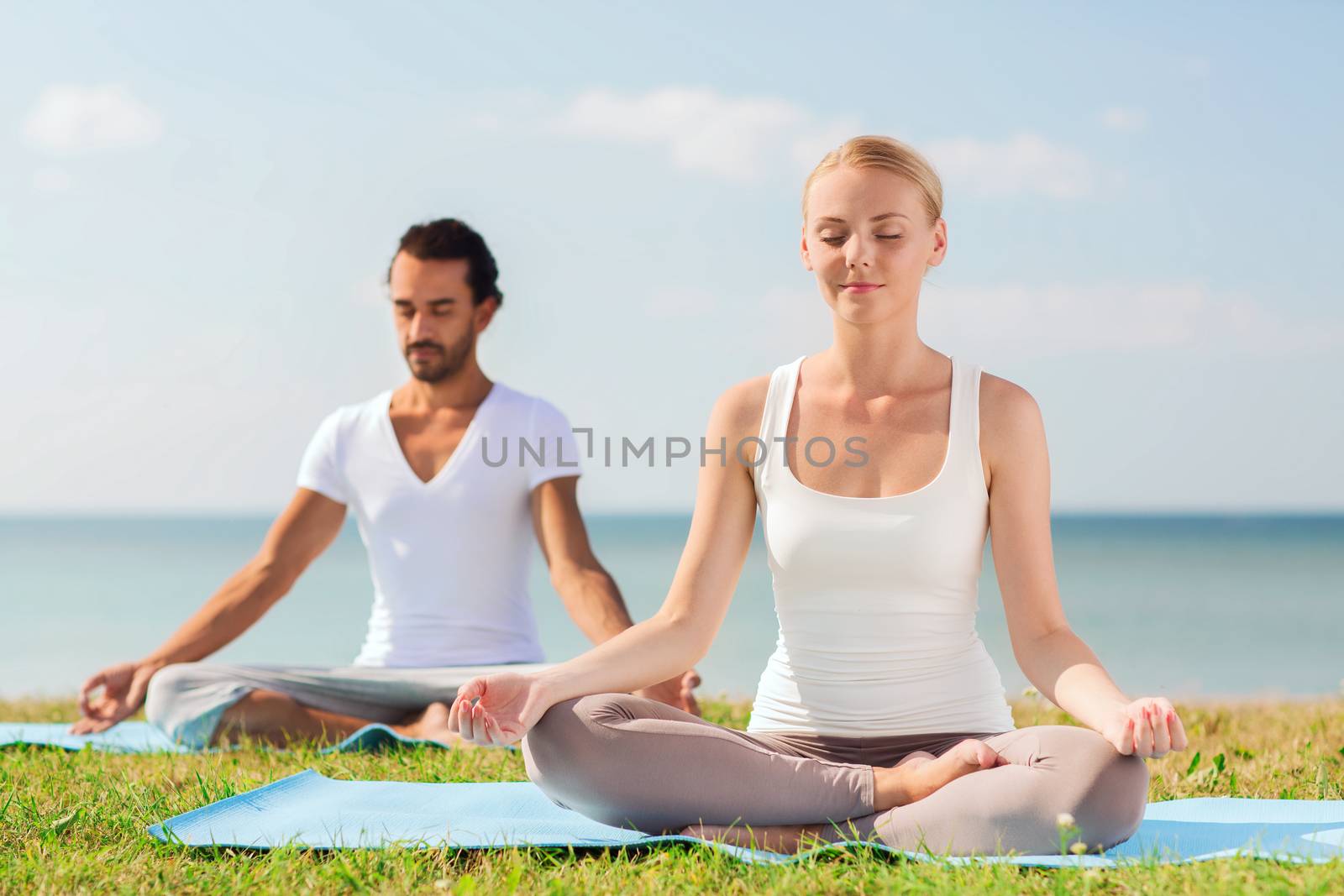 fitness, sport, friendship and lifestyle concept - smiling couple making yoga exercises sitting on mats outdoors