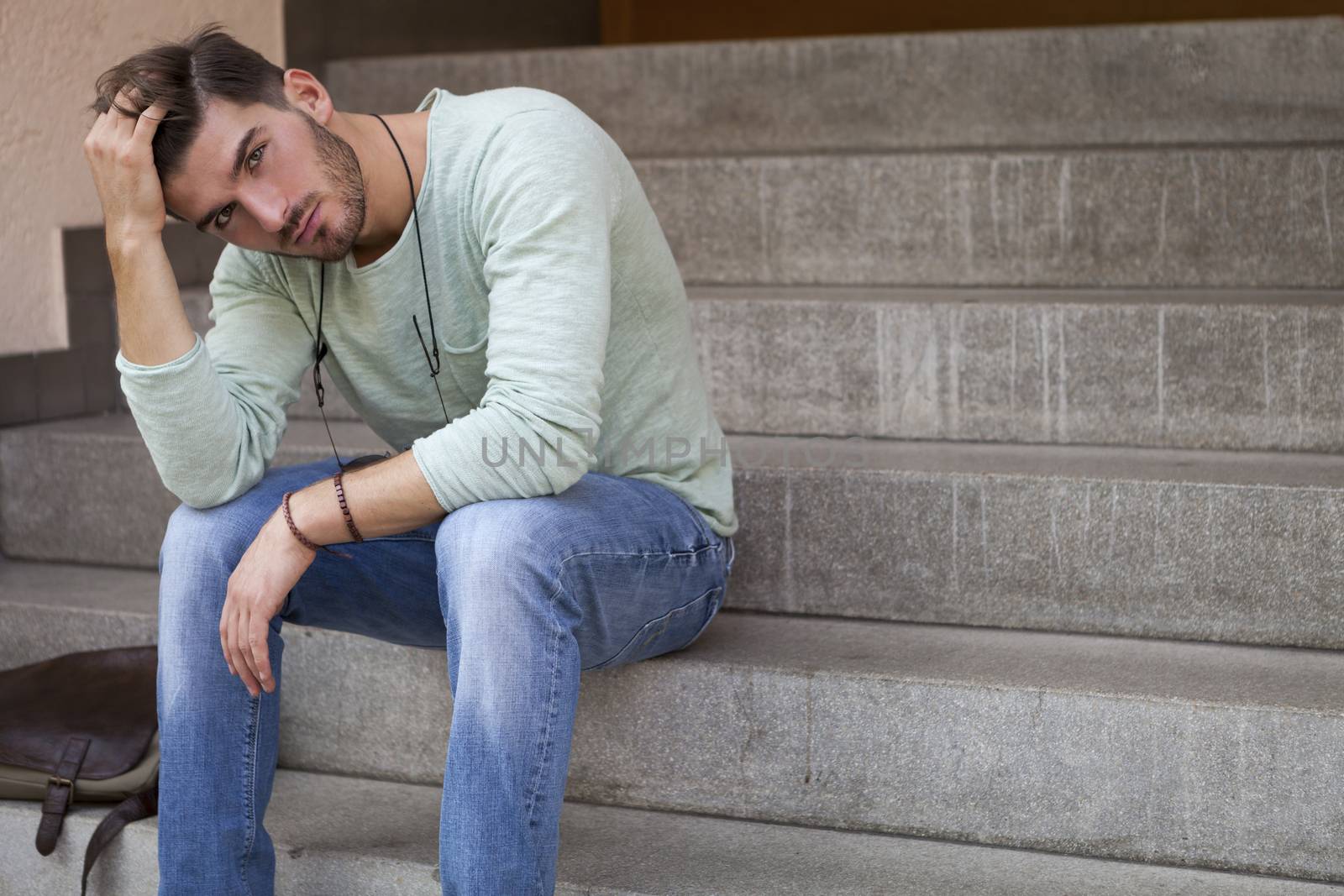 Casual  handsome young man with a beard sitting on cement steps with his head resting on his hand and a serious dejected expression