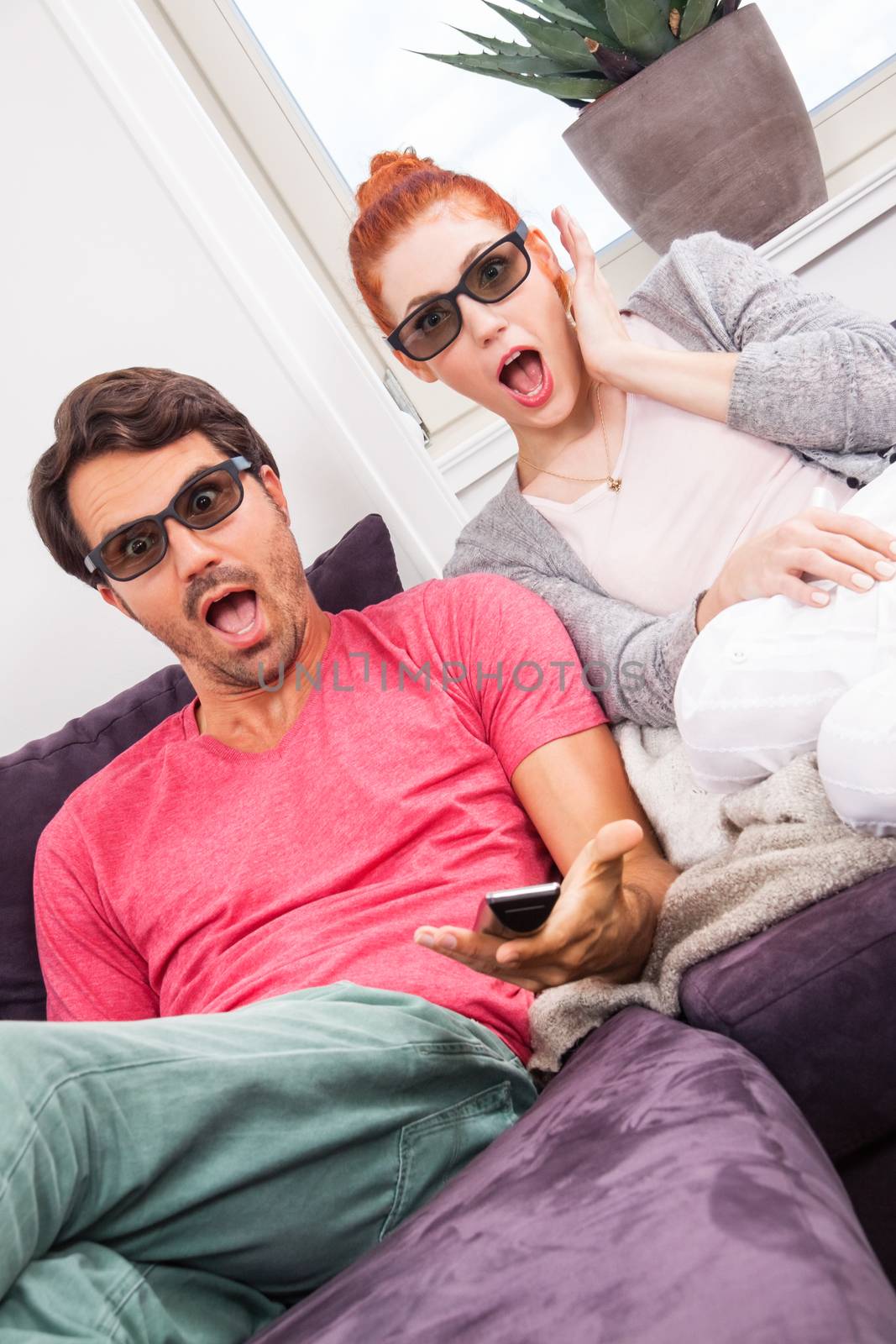 Young Couple Wearing 3D Glasses, Sitting on the Couch in the Living Room, Watching a Movie and Showing Shocked Faces.