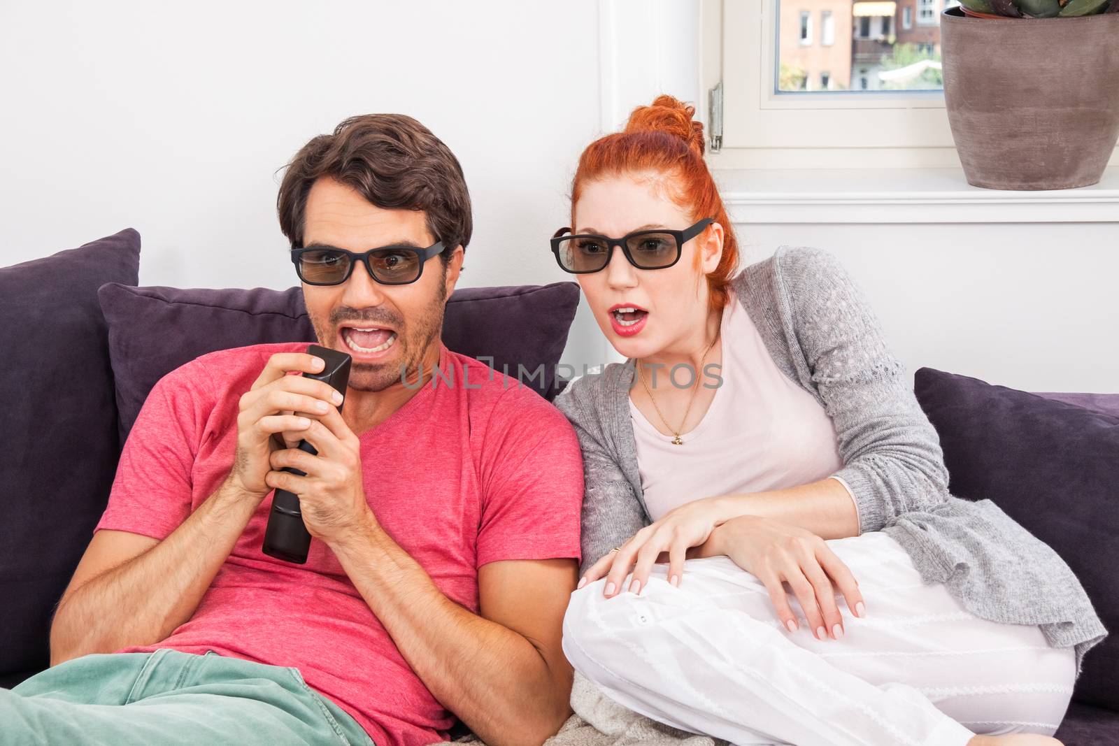 Young Couple Wearing 3D Glasses, Sitting on the Couch in the Living Room, Watching a Movie and Showing Shocked Faces.