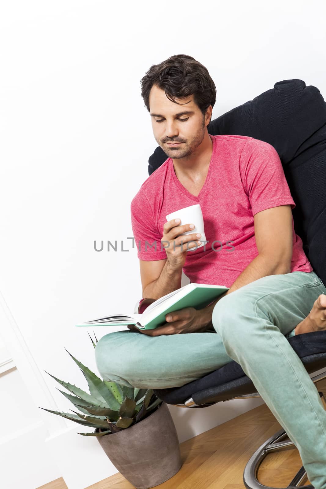 Young Man in Casual Clothing Sitting on Black Chair While Reading a Book and Holding a Glass of Drink.