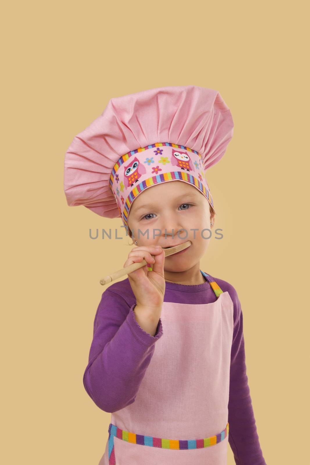 Cute baby girl chef cook looking into camera, tasting food from wooden scoop isolated. Baking and cooking. 