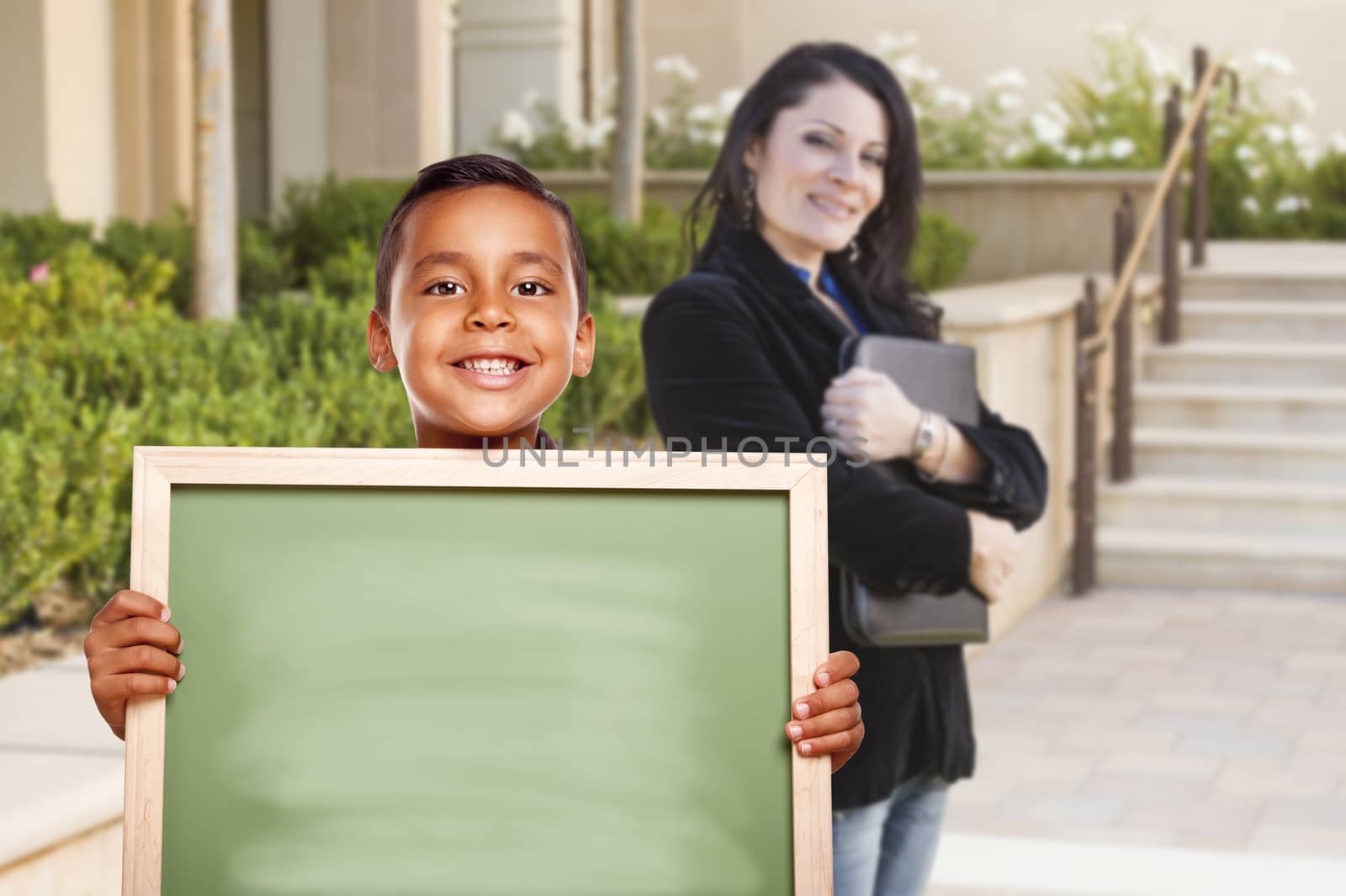 Happy Hispanic Boy Holding Blank Chalk Board on Campus As Teacher Looks on From Behind.