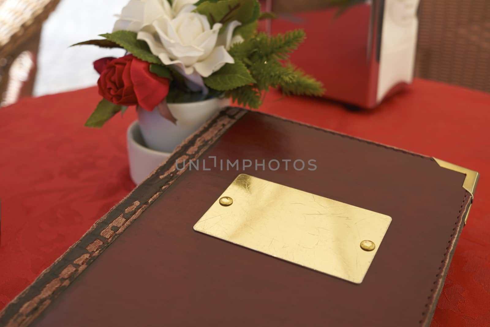 Detail of leather bound menu with blank name plate on red table cloth. There's a small flower vase on the side with red and white roses.