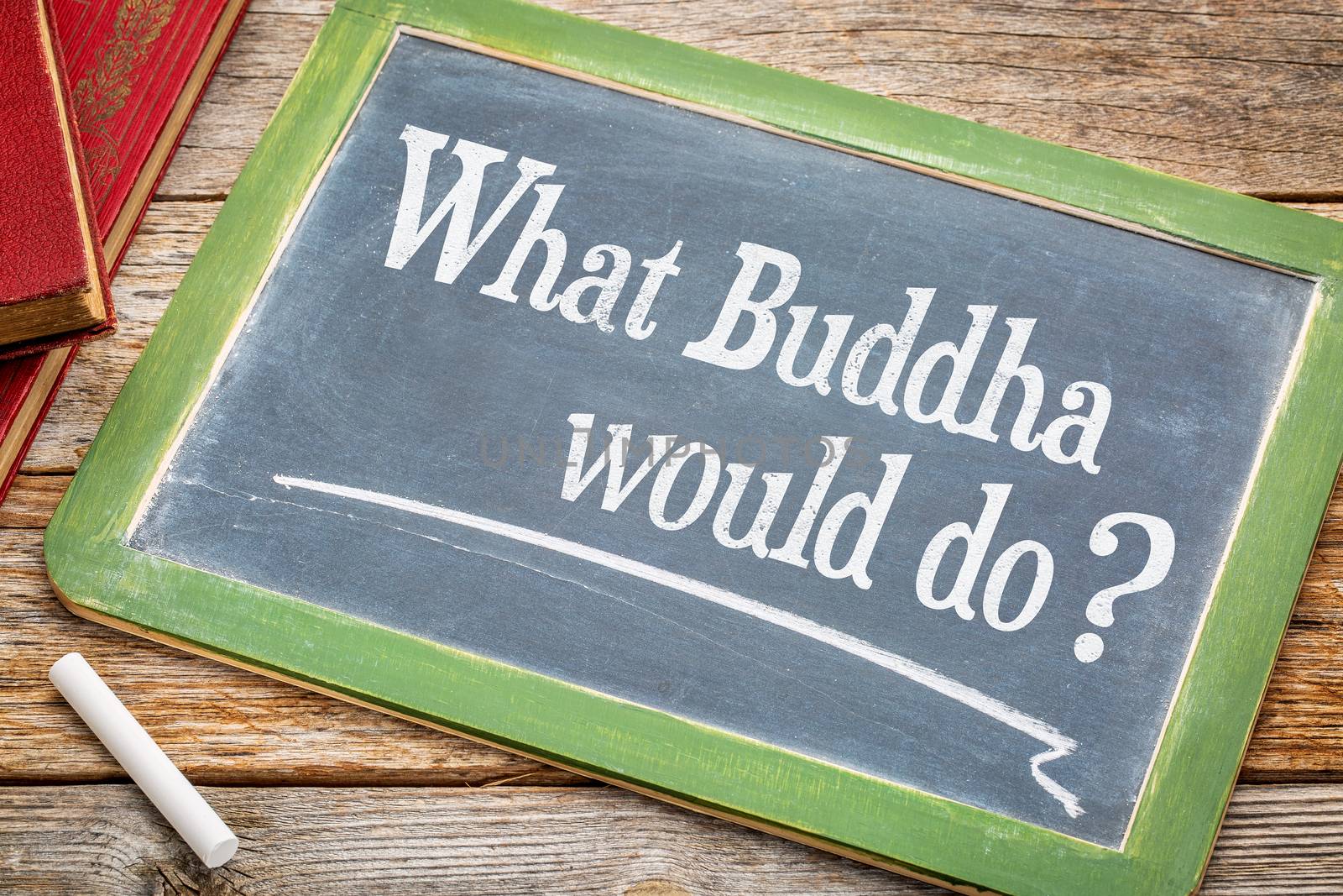 What Buddha would? An inspirational question on a slate blackboard with a white chalk and a stack of books against rustic wooden table