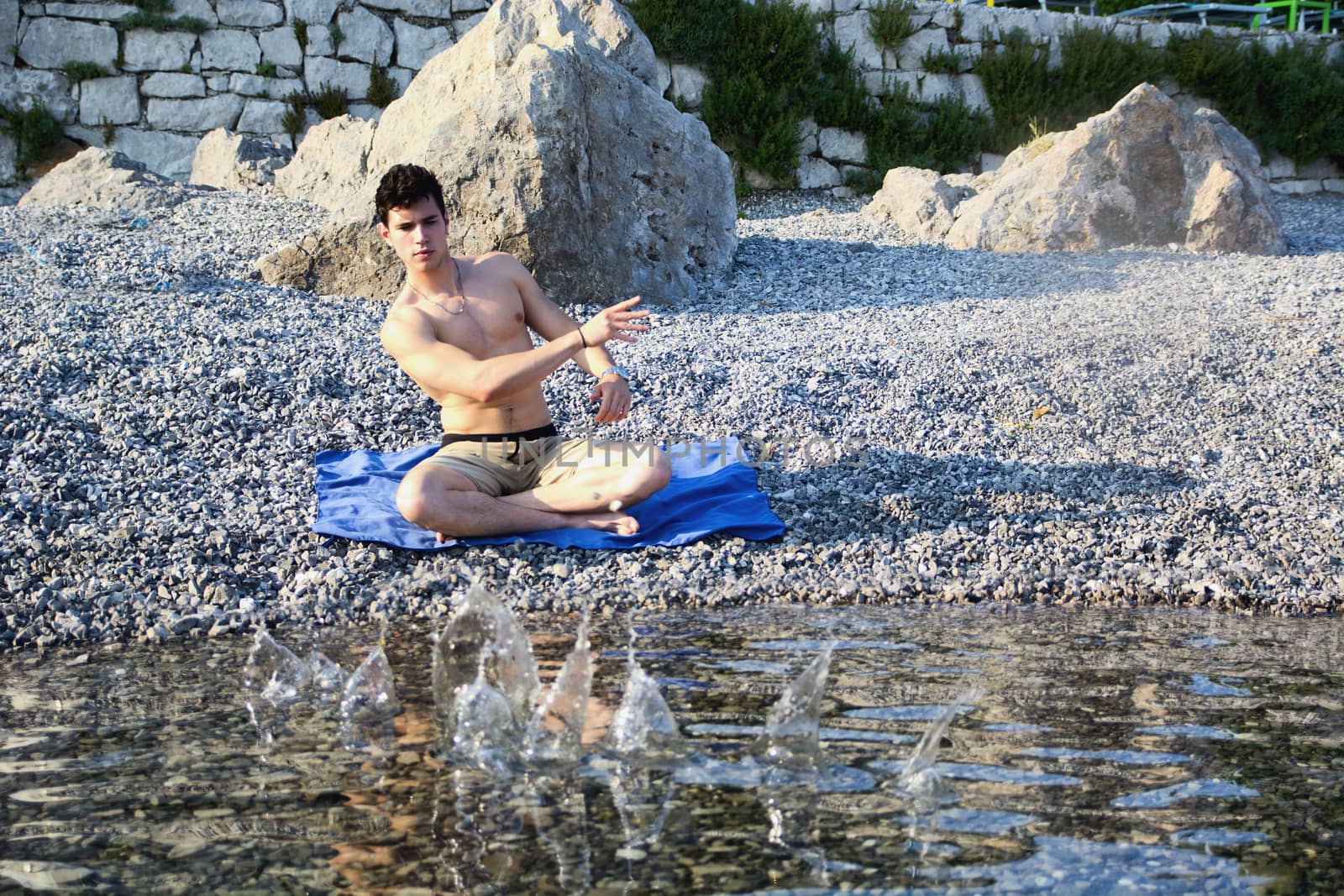 Young Shirtless Man Sitting Cross Legged on Blue Blanket on Rocky Beach and Skipping Stones Across Water Surface from Shore