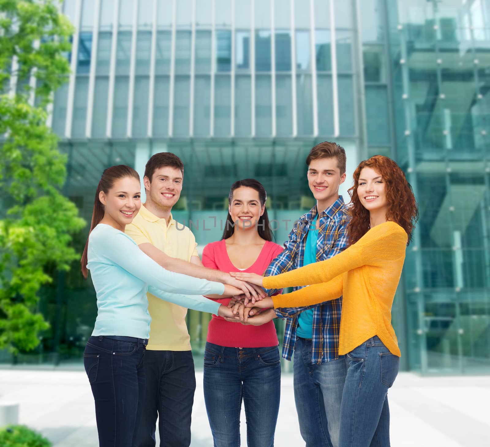 friendship, education, business, gesture and people concept - group of smiling teenagers with hands on top of each other over campus background