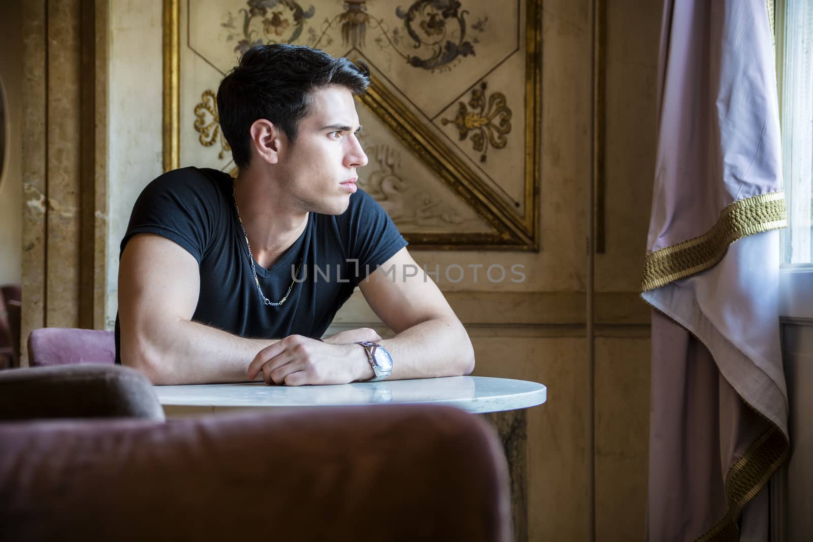 Portrait of Serious Attractive Young Man Sitting at Table Leaning on it and Looking Away, in Room Decorated with Gold Framed Artwork