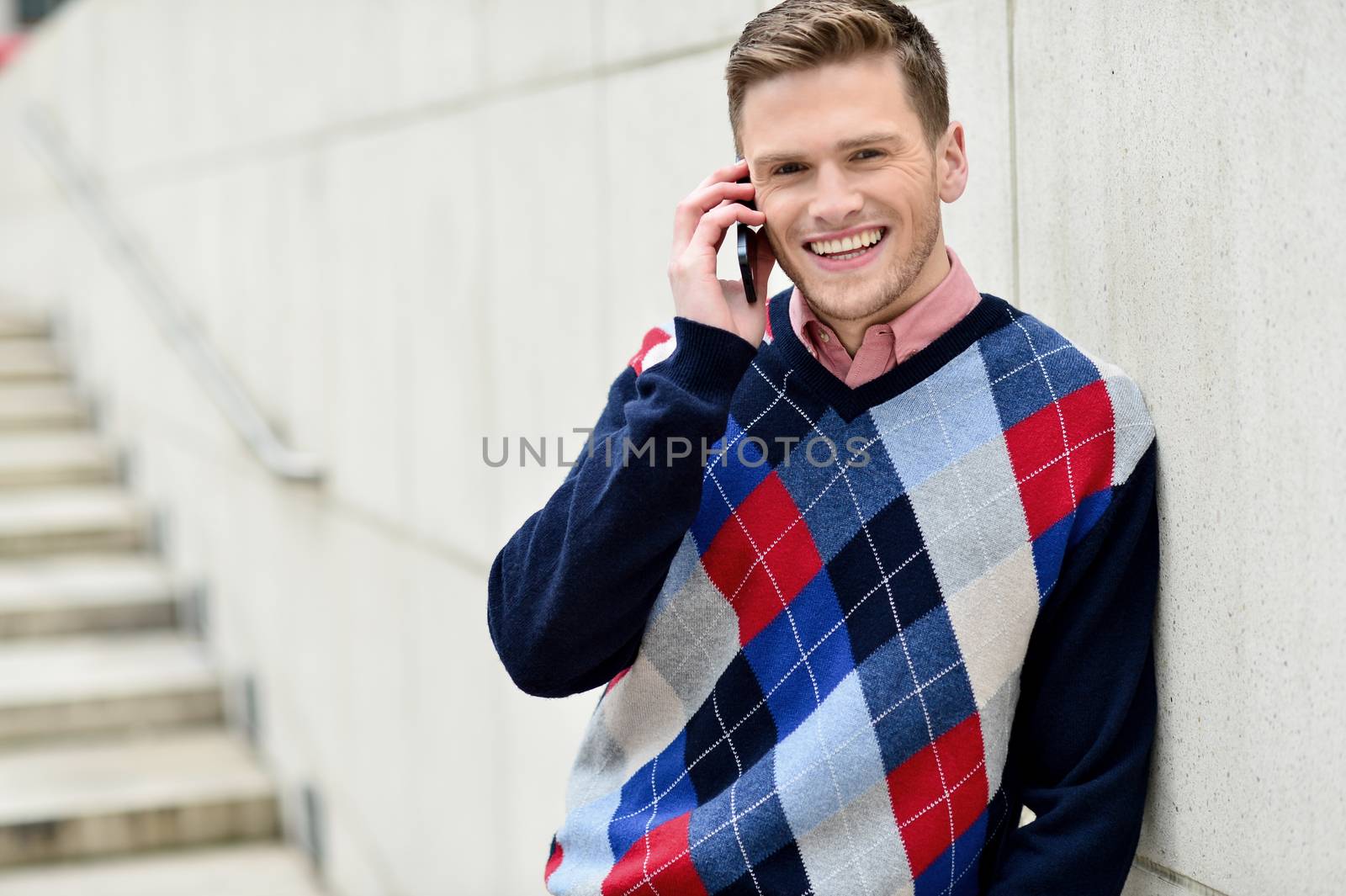 Casual man talking on cell phone, leaning on wall. 