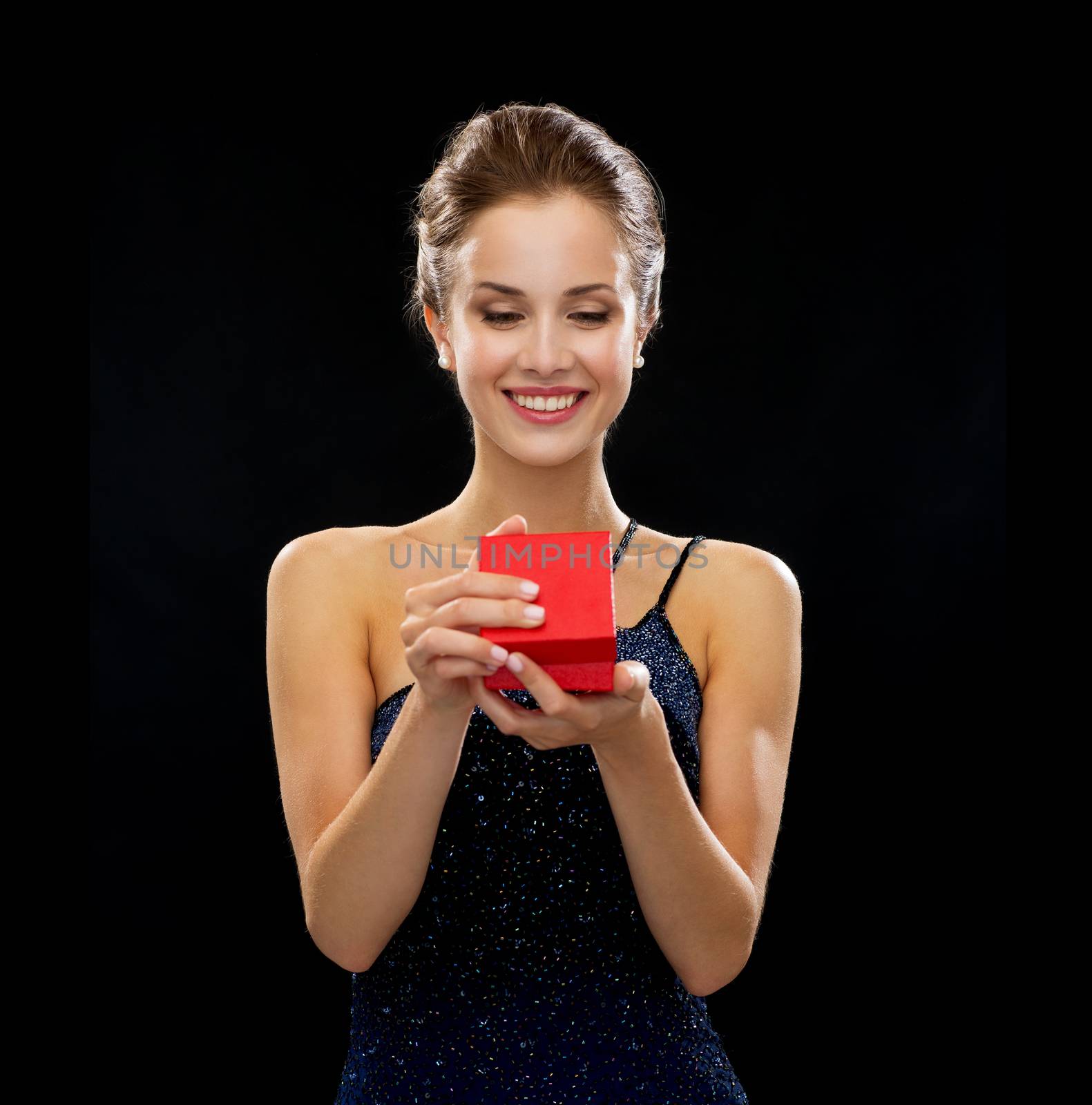 holidays, presents, luxury and happiness concept - smiling woman in dress holding red gift box over black background