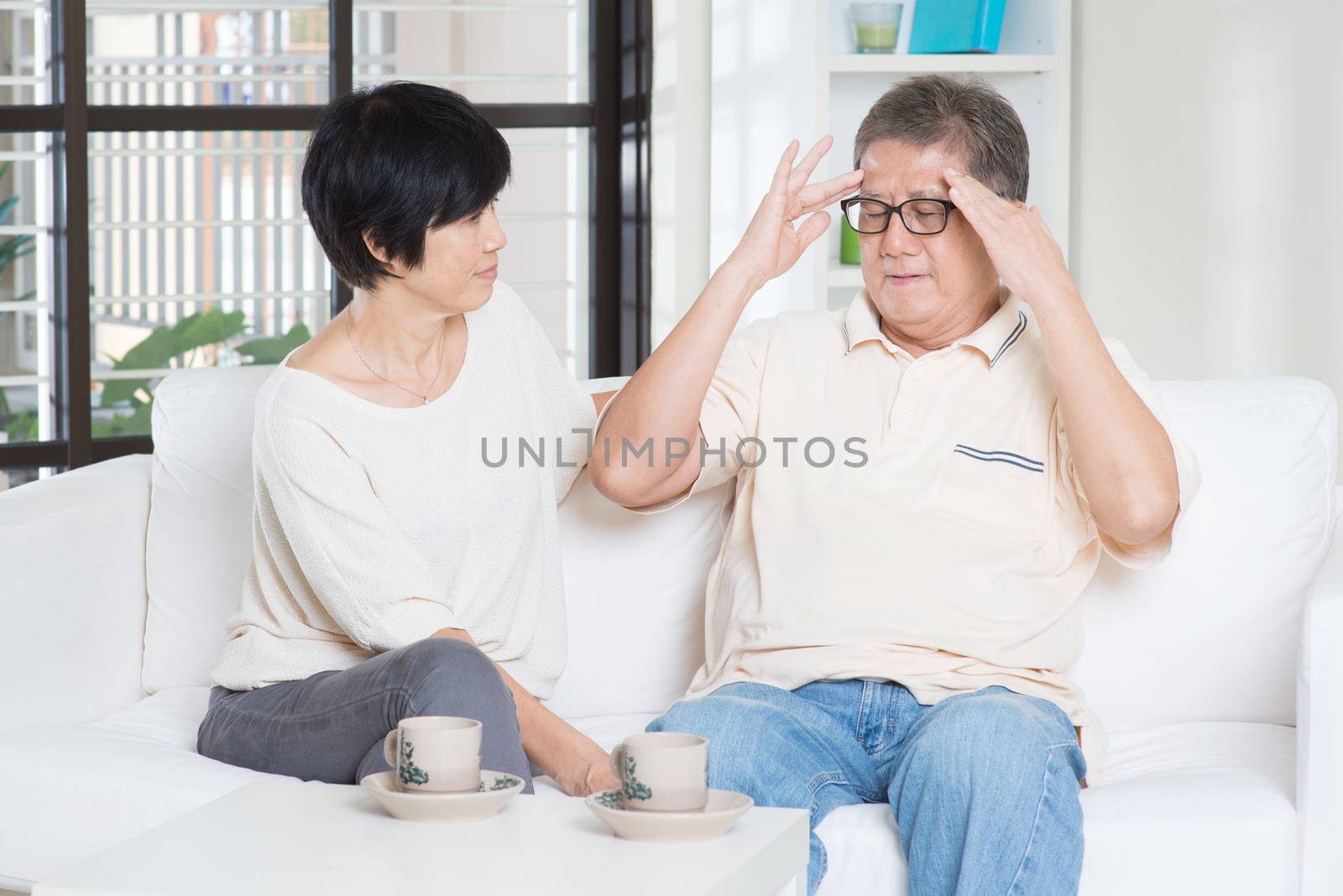 Asian old man headache, sitting on sofa with wife at home. Chinese family, senior retiree indoors living lifestyle.