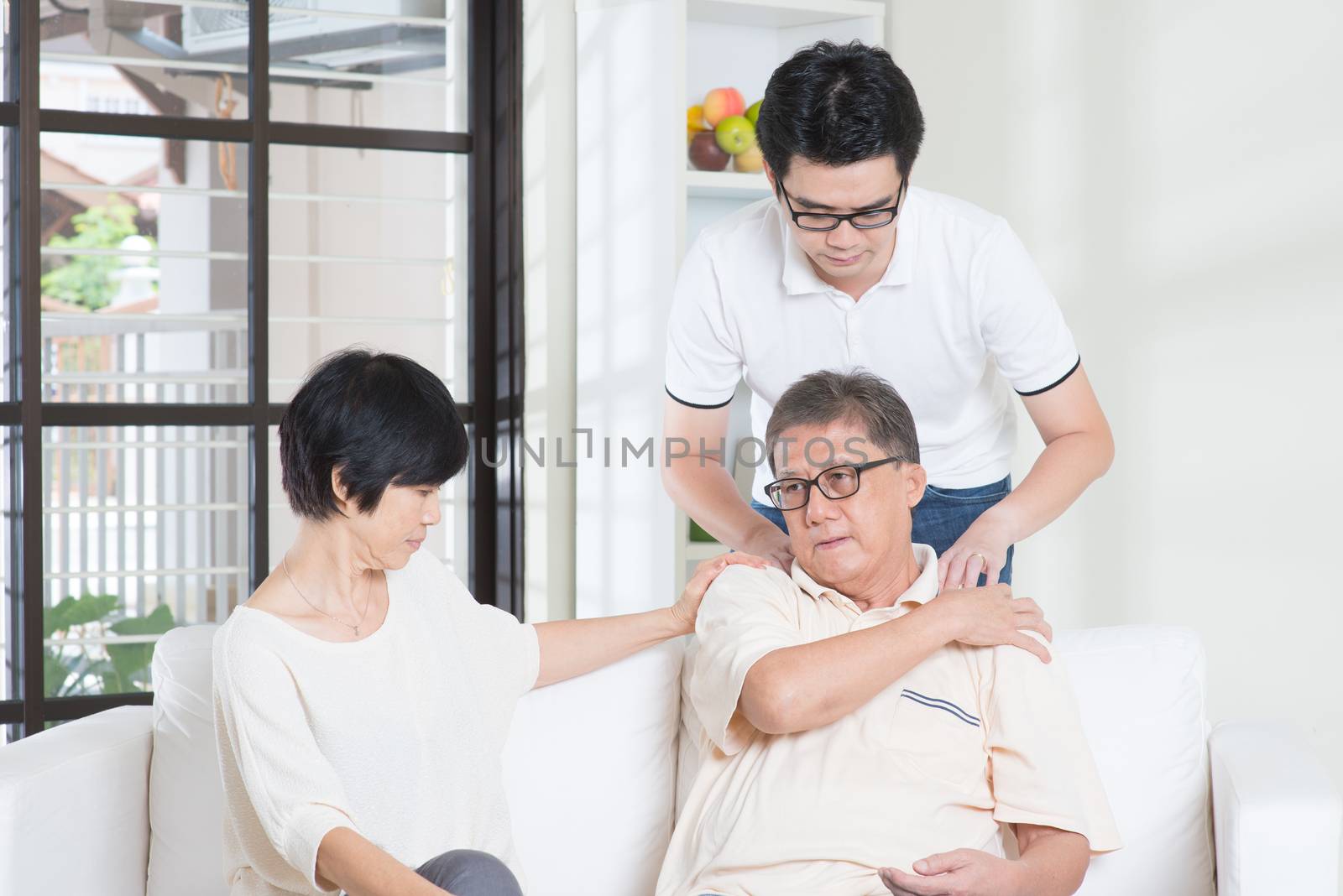 Asian old man shoulder pain, sitting on sofa with wife, son massaging father shoulder. Chinese family, senior retiree indoors living lifestyle at home.