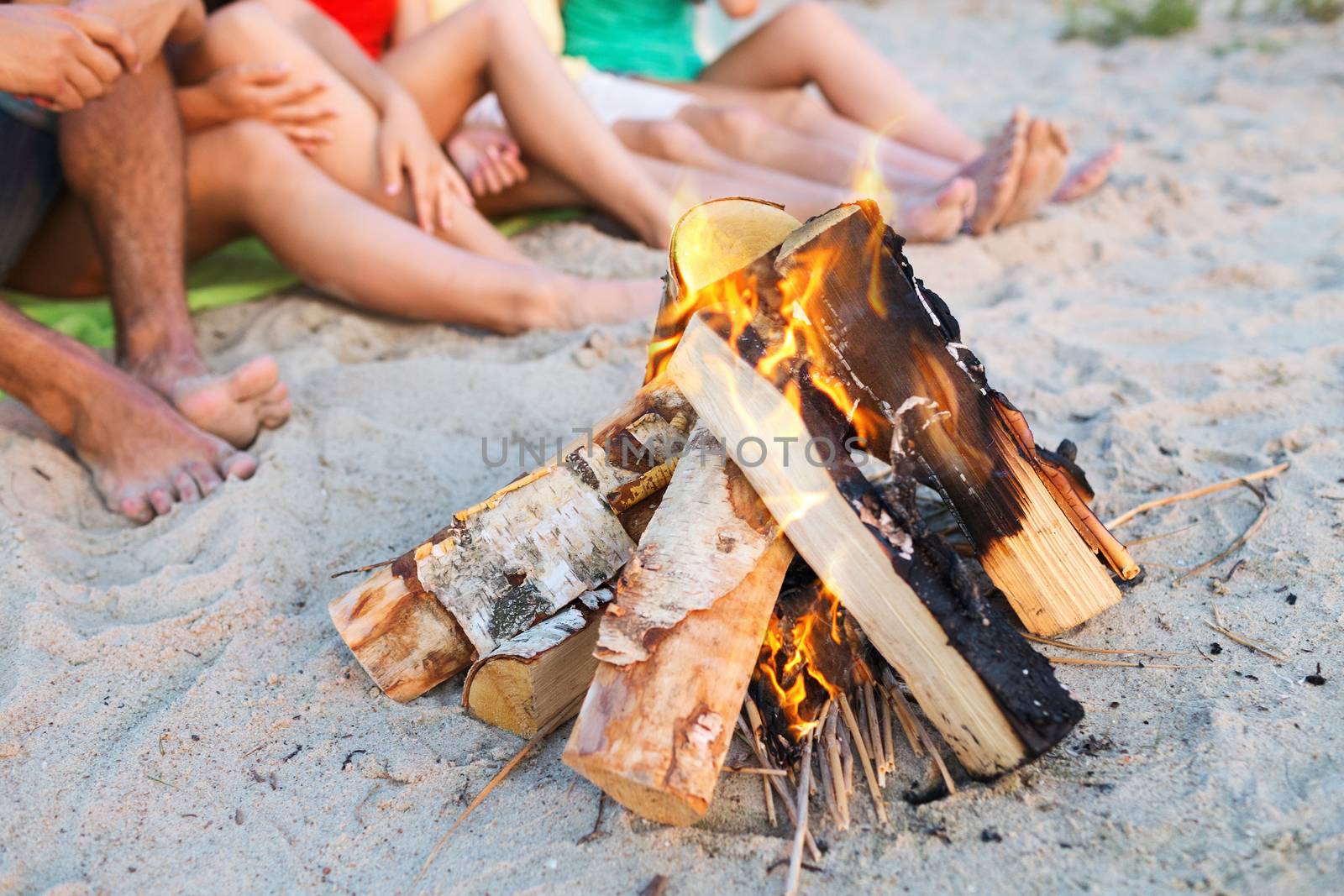 friendship, happiness, summer vacation, holidays and people concept - close up of friends sitting near fire on beach