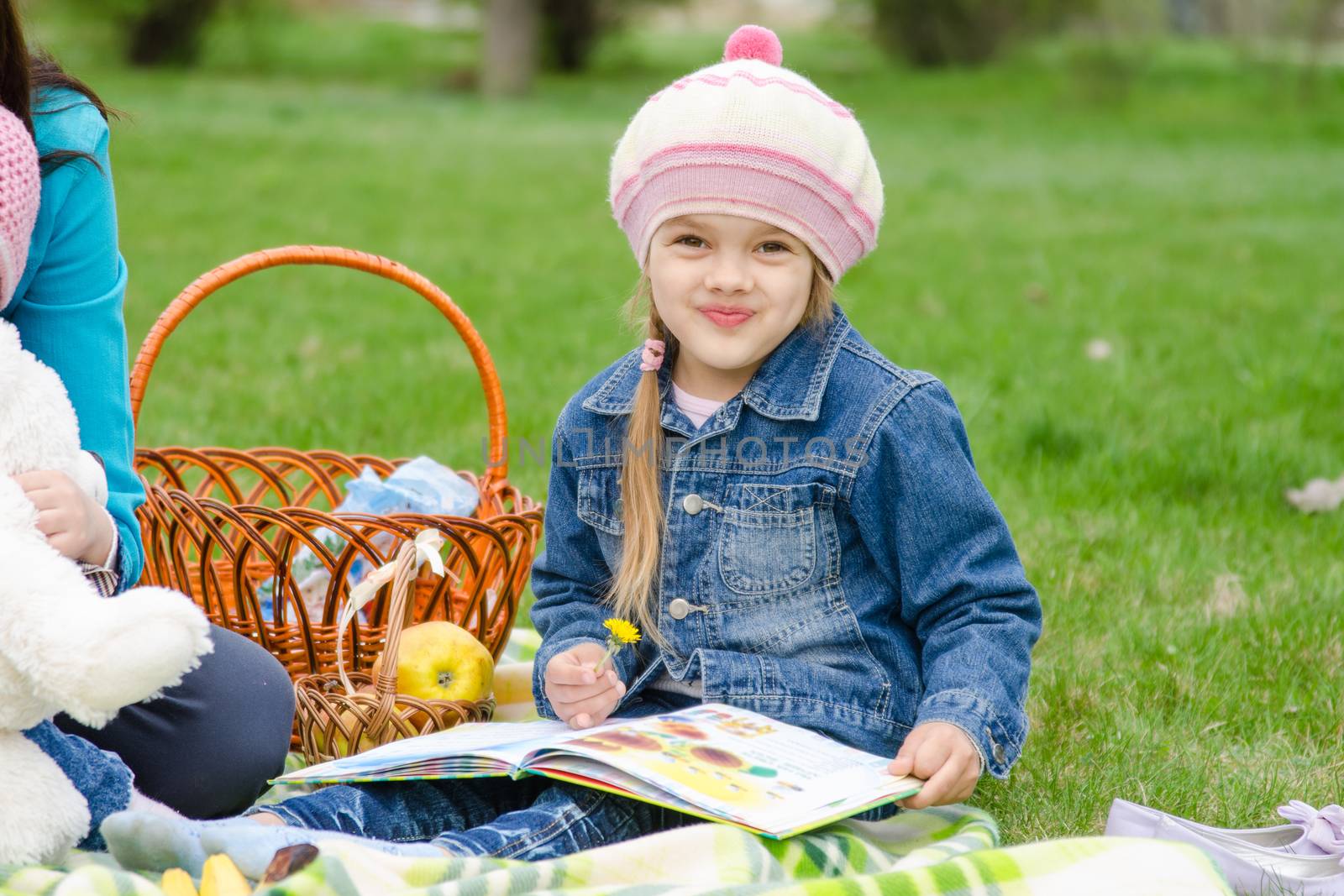 A girl of five years in a denim jacket on a green lawn at a picnic with a book in their hands