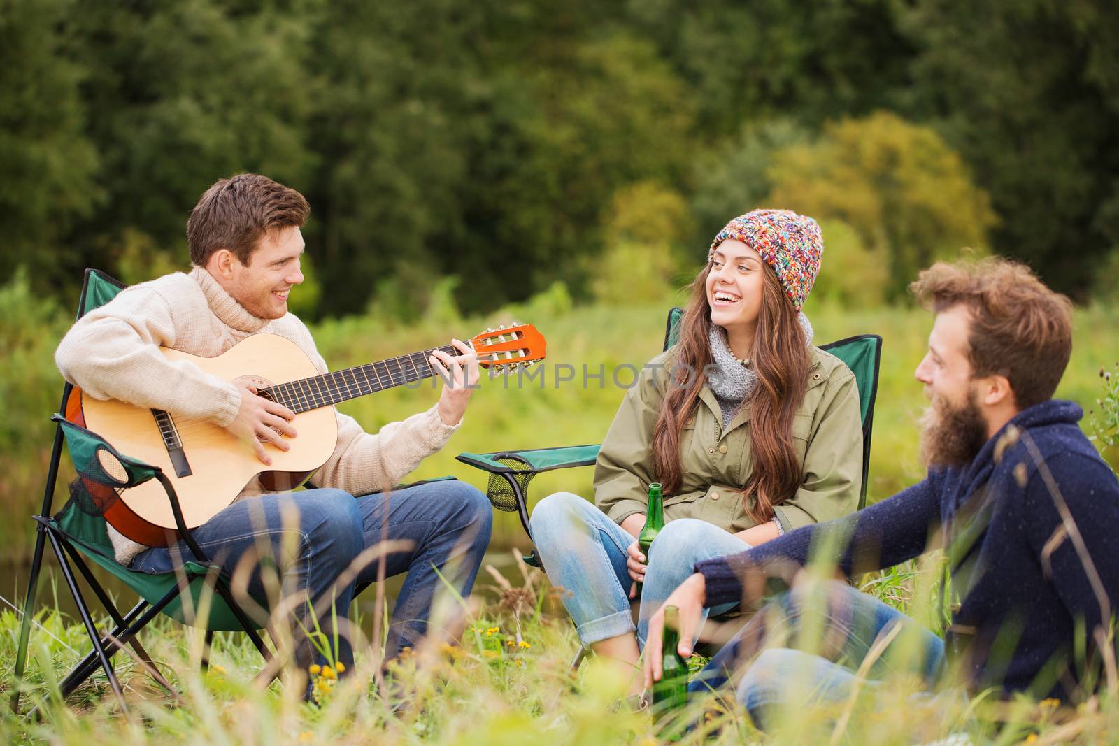 adventure, travel, tourism and people concept - group of smiling friends with guitar sitting on chairs in camping