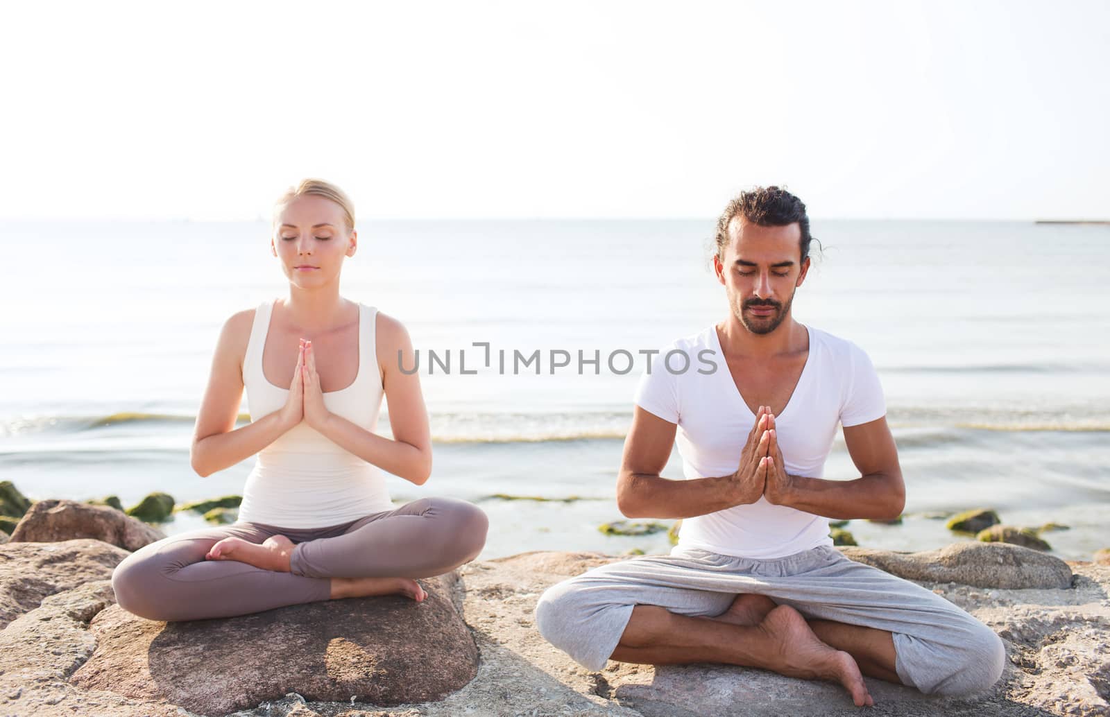 fitness, sport, friendship and lifestyle concept - smiling couple making yoga exercises sitting on sand outdoors