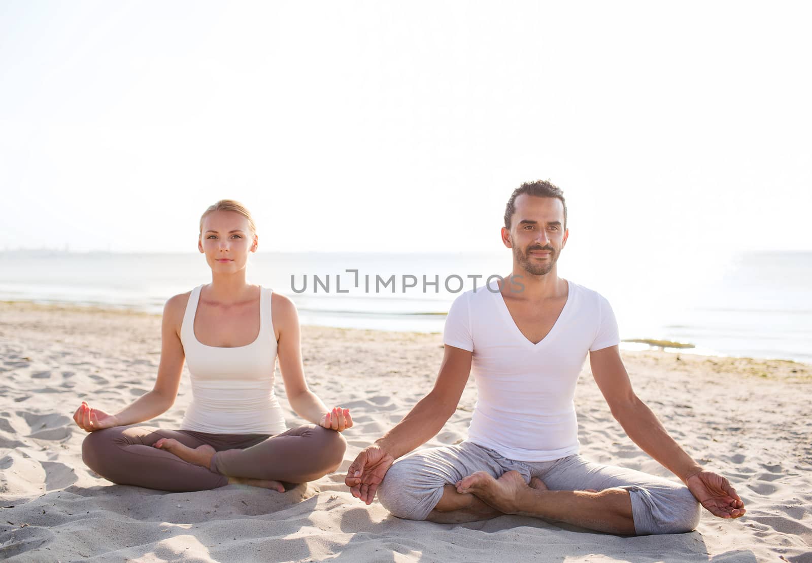 fitness, sport, friendship and lifestyle concept - smiling couple making yoga exercises sitting on sand outdoors