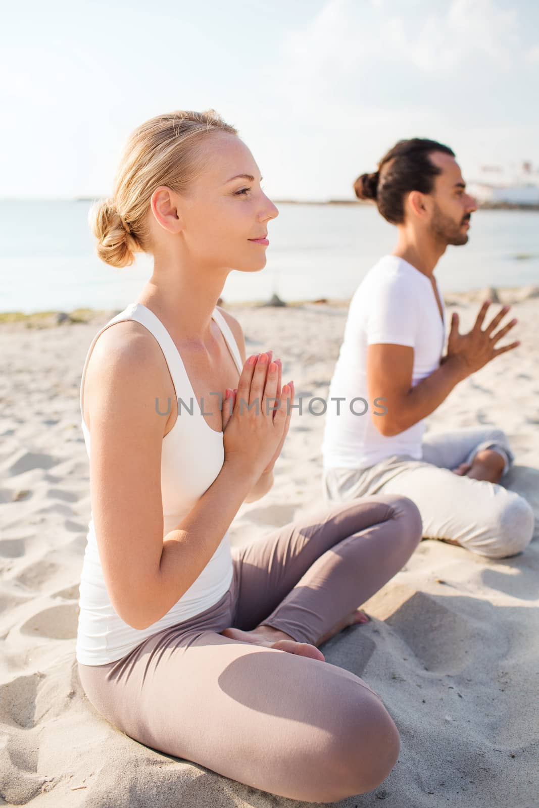 fitness, sport, friendship and lifestyle concept - smiling couple making yoga exercises sitting on sand outdoors