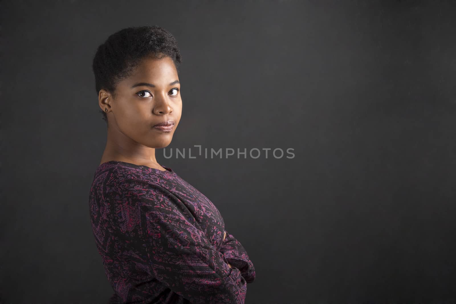 South African or African American woman teacher or student with arms folded on chalk black board background by alistaircotton