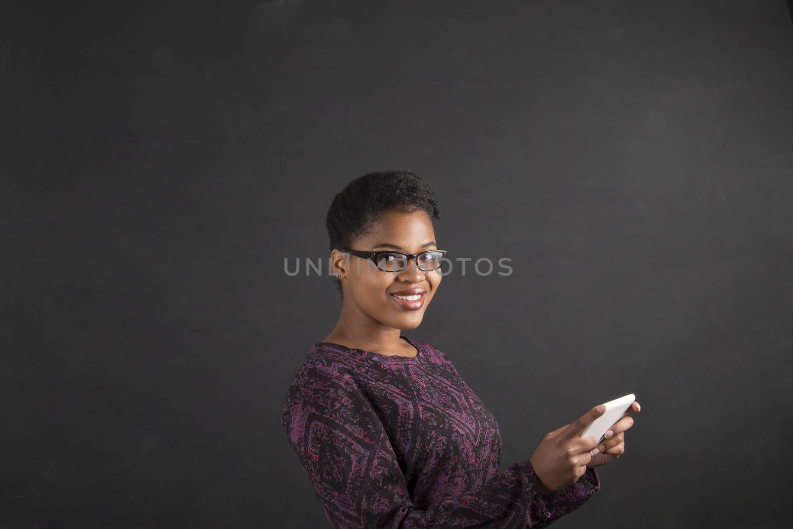 South African or African American black woman teacher or student holding a tablet standing against a chalk blackboard background inside