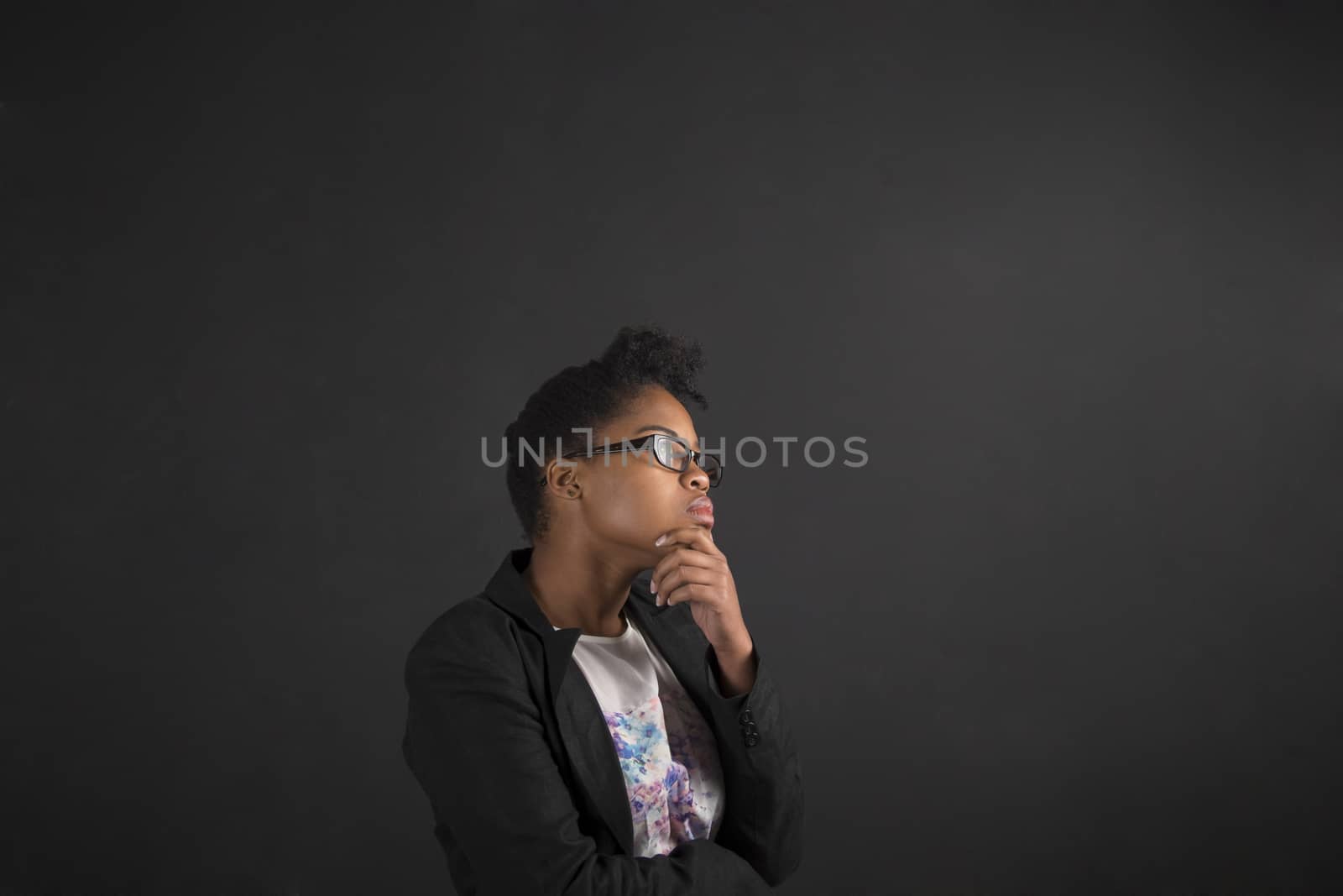 South African or African American black woman teacher or student with her hand on her chin whilst thinking standing against a chalk blackboard background inside