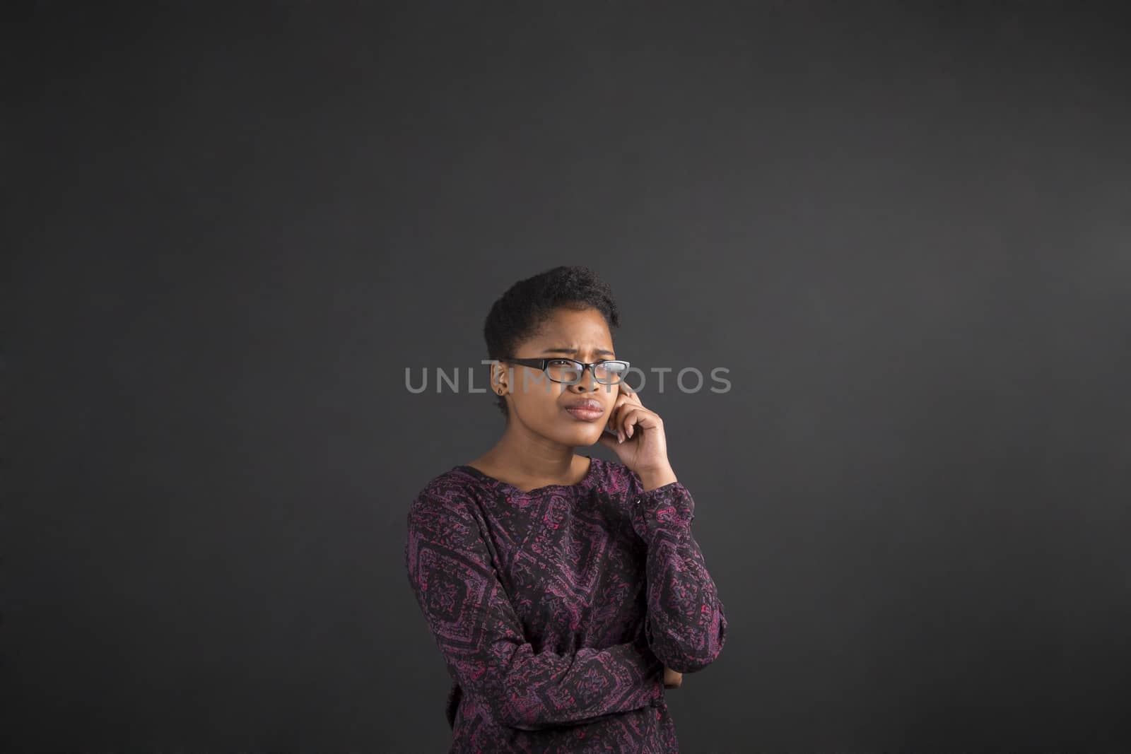 South African or African American black woman teacher or student with her hand on her chin whilst thinking standing against a chalk blackboard background inside
