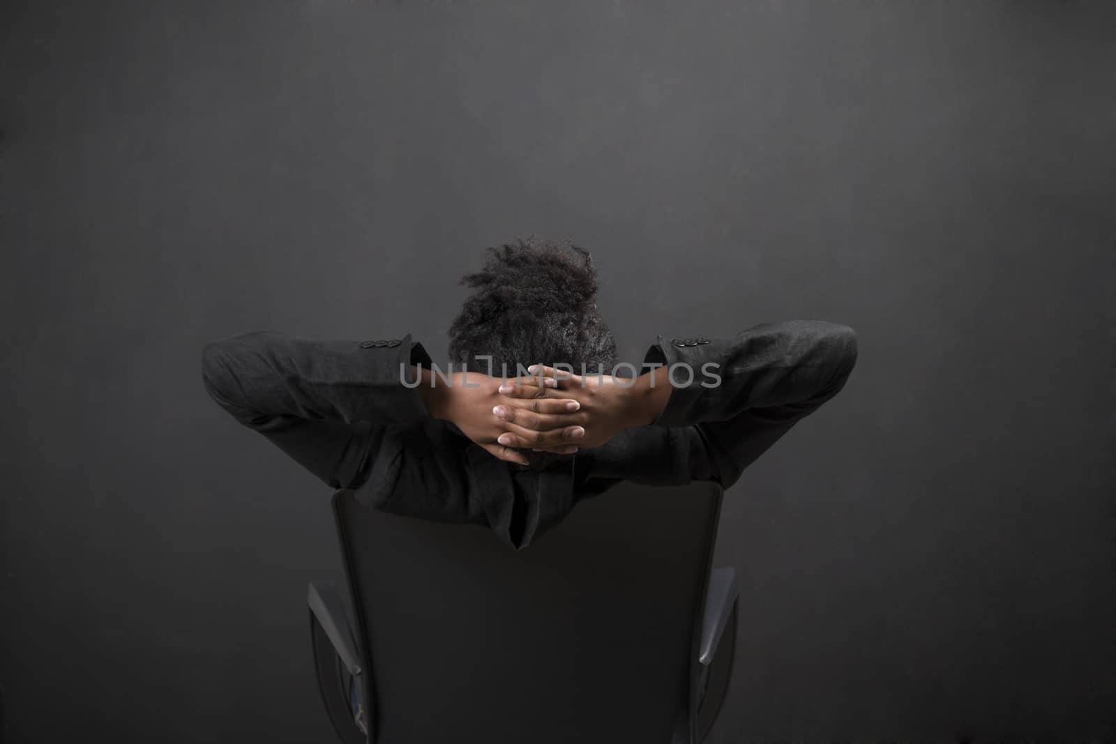 South African or African American black woman teacher or student sitting on a chair with her arms behind her head against a chalk blackboard background inside