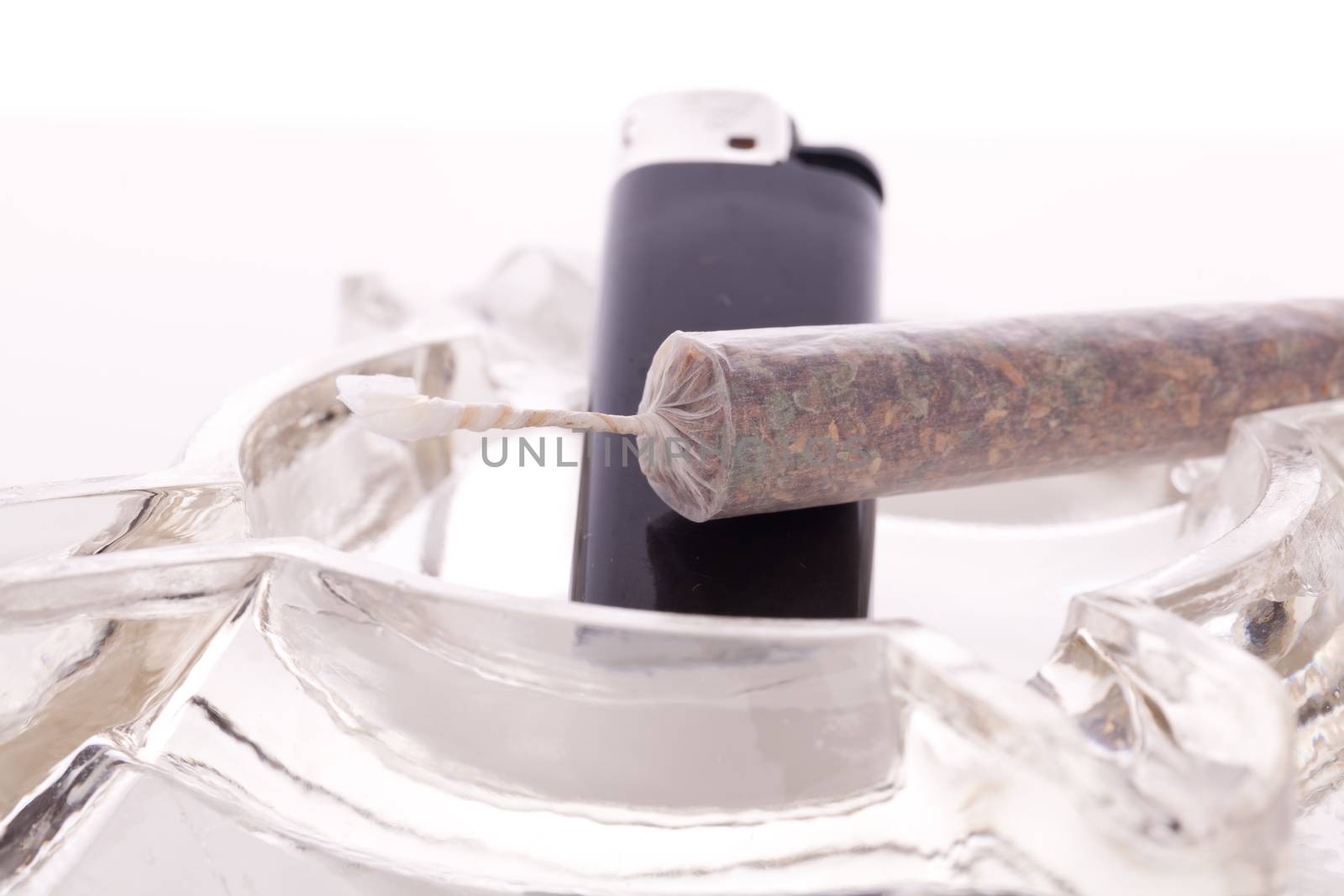 Close up of marijuana joint made with translucent rolling papers, plastic baggy of dried marijuana, black lighter and pipe on white background