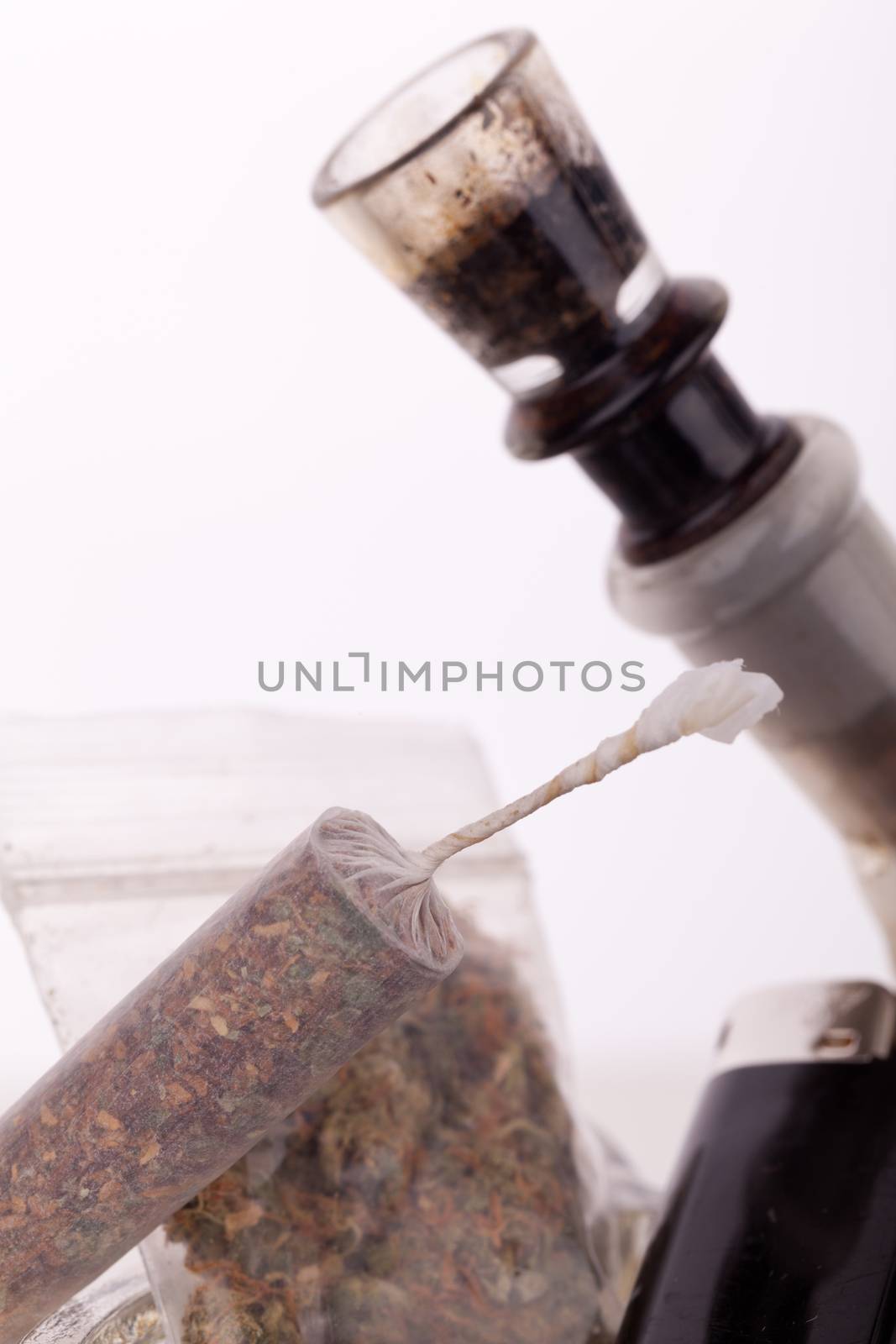 Close up of marijuana joint made with translucent rolling papers, plastic baggy of dried marijuana, black lighter and pipe on white background