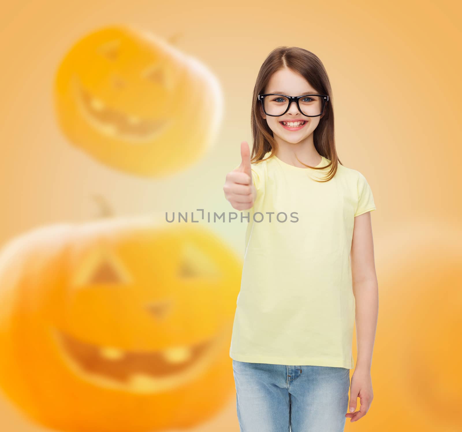 gesture, education, holidays and people concept - smiling little girl in glasses showing thumbs up over halloween pumpkins background