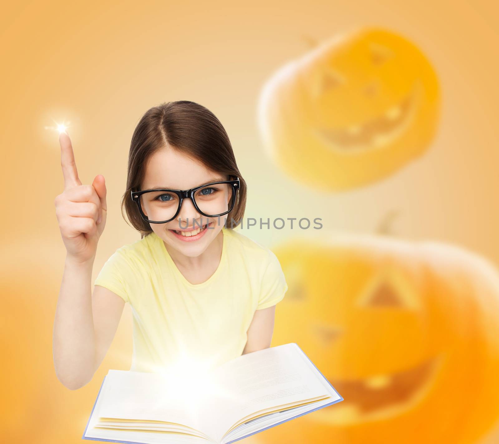 education, holidays, childhood, vision and people concept - smiling little girl in glasses with magic book over halloween pumpkins background