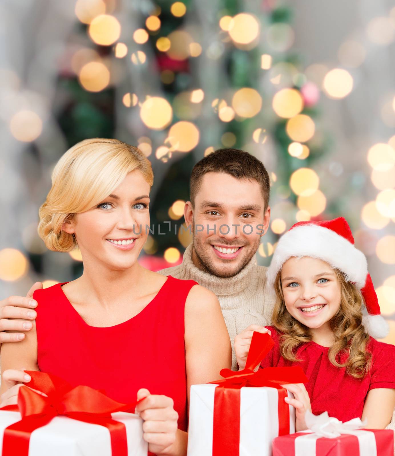 christmas, holidays, family and people concept - happy mother, father and little girl in santa helper hat with gift boxes over over living room and christmas tree background
