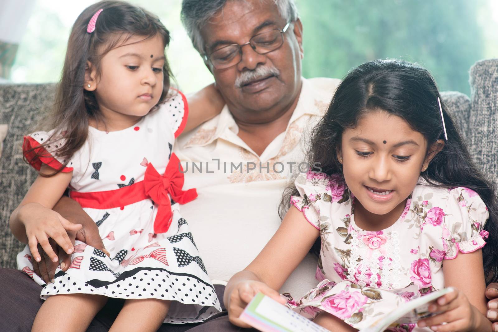 Happy Indian family at home. Asian grandfather and granddaughters reading story book. Grandparent and grandchildren indoor lifestyle.