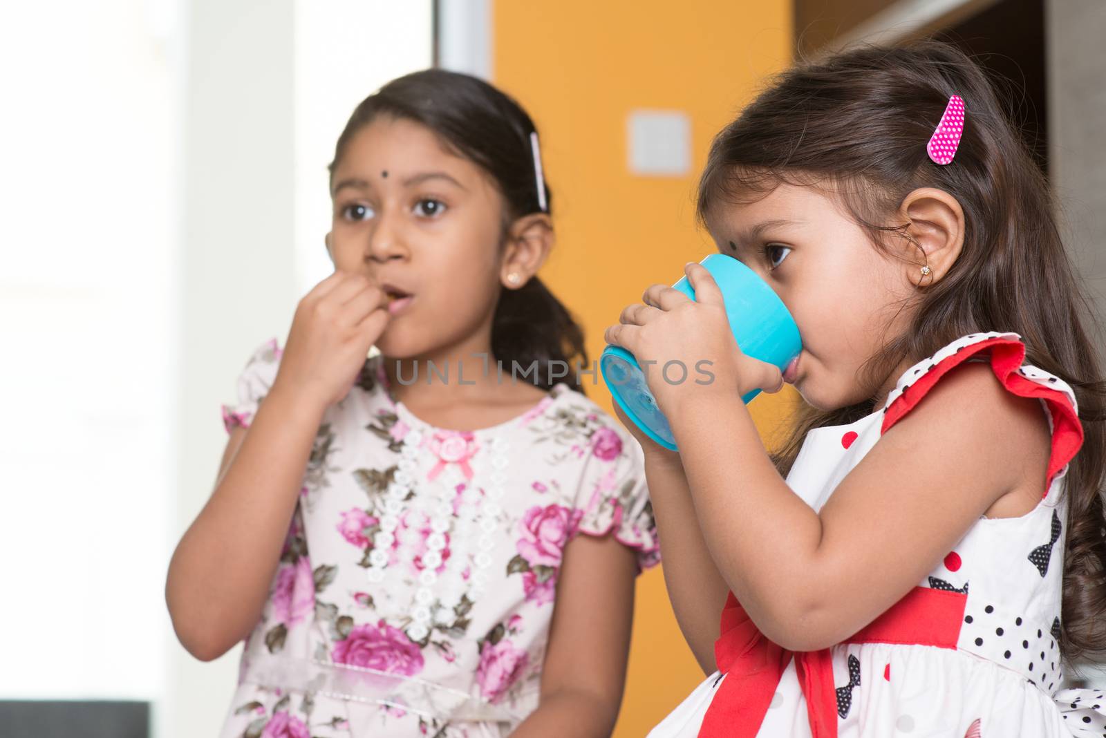 Two cute Indian girls eating food and drinking water. Asian sibling or children enjoying tea time food, living lifestyle at home.