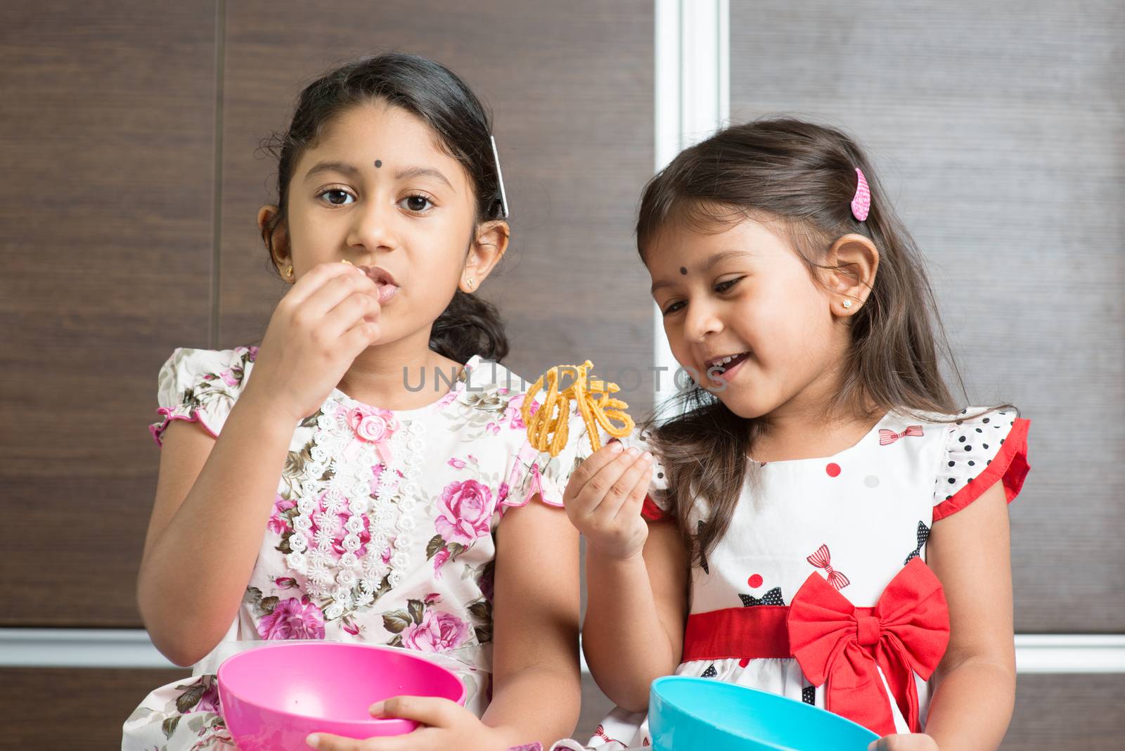 Two cute Indian girls eating food. Asian sibling or children enjoying traditional snack murukku, living lifestyle at home.