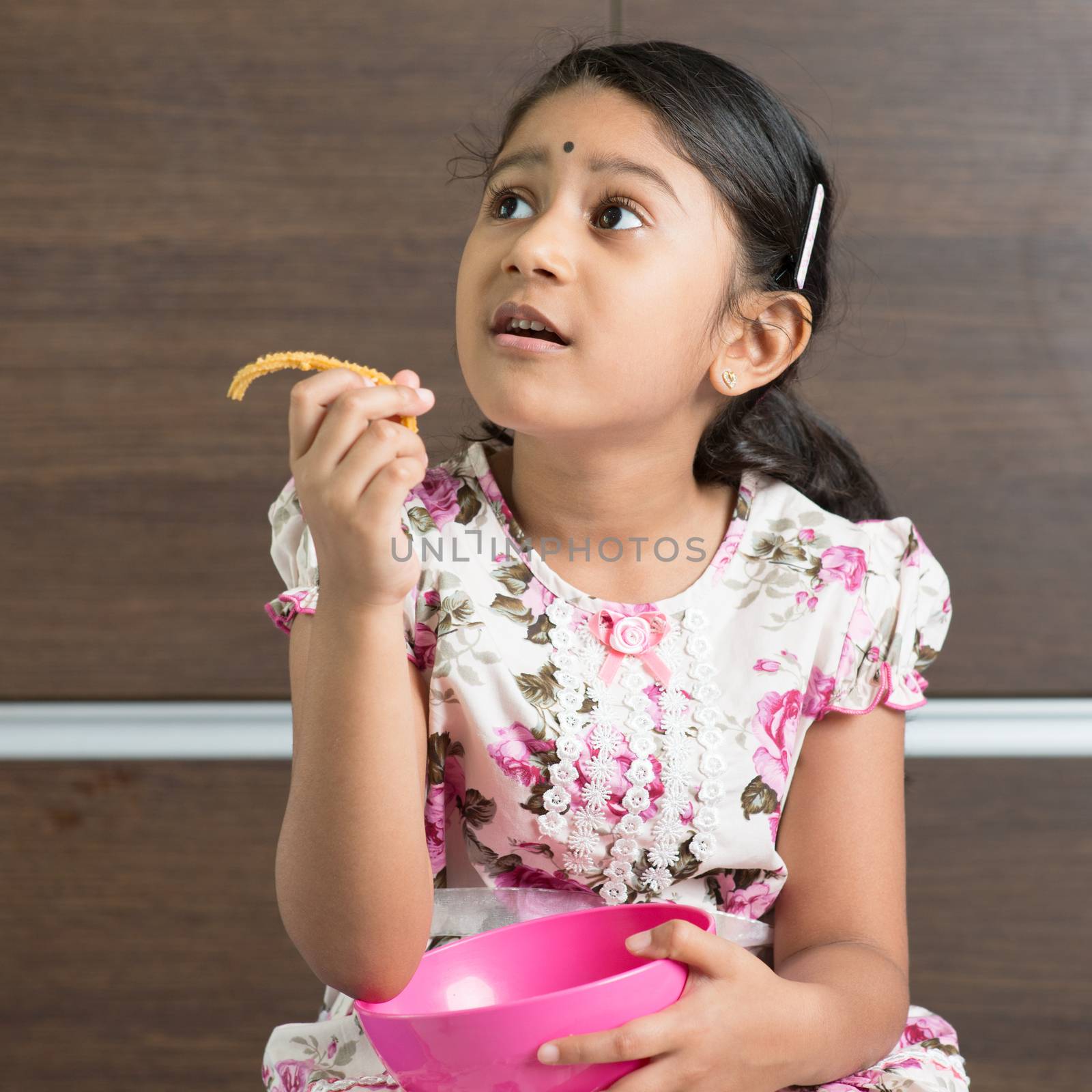 Cute Indian girl eating traditional snack murukku. Asian child enjoying food, living lifestyle at home.