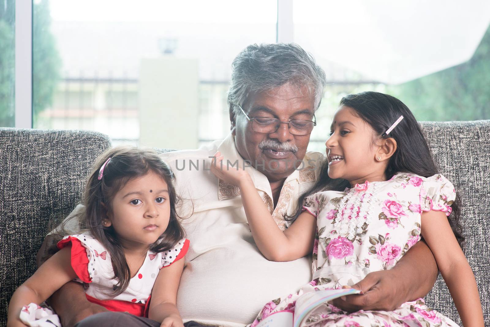 Grandparent and grandchildren reading story book together. Happy Indian family at home. Asian grandfather and granddaughters indoor lifestyle.