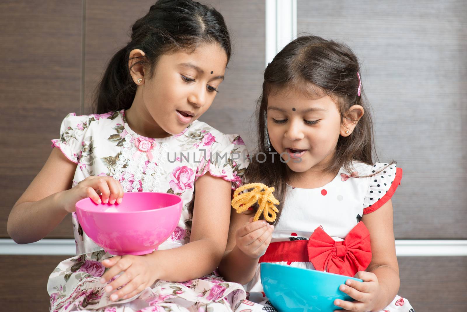 Two cute Indian girls eating traditional snack murukku. Asian sibling or children enjoying food, living lifestyle at home.