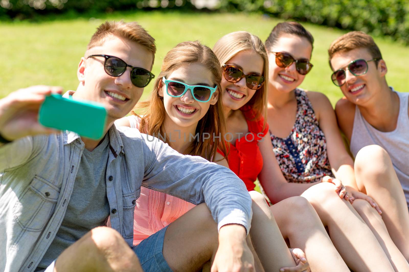 smiling friends with smartphone sitting on grass by dolgachov