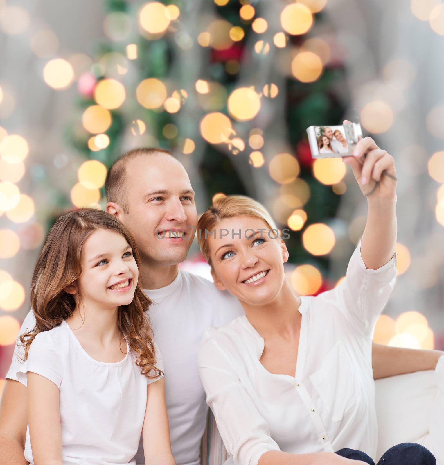 family, holidays, technology and people - smiling mother, father and little girl making selfie with camera over christmas tree lights background