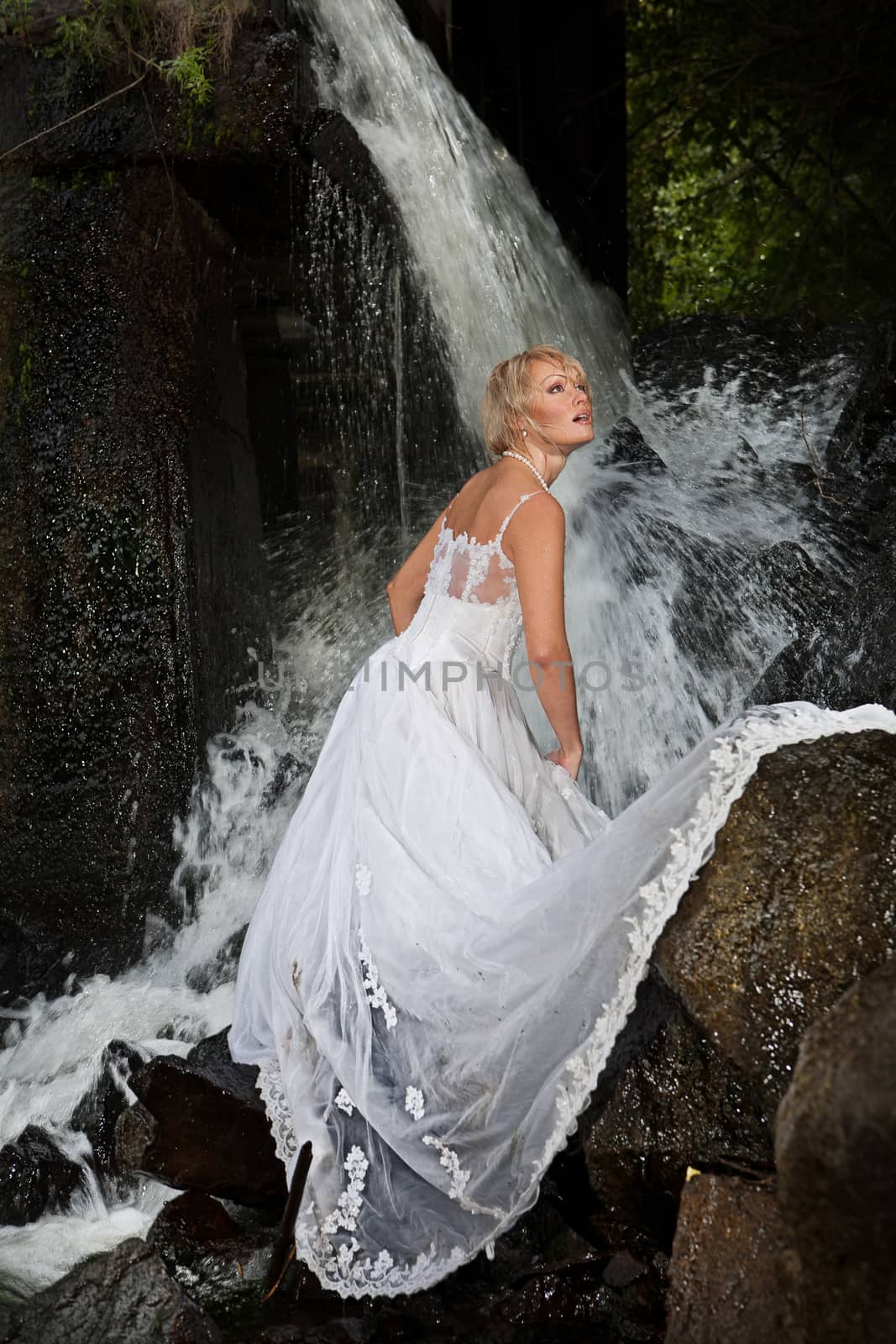 Young blonde woman in a white wedding dress near the waterfall